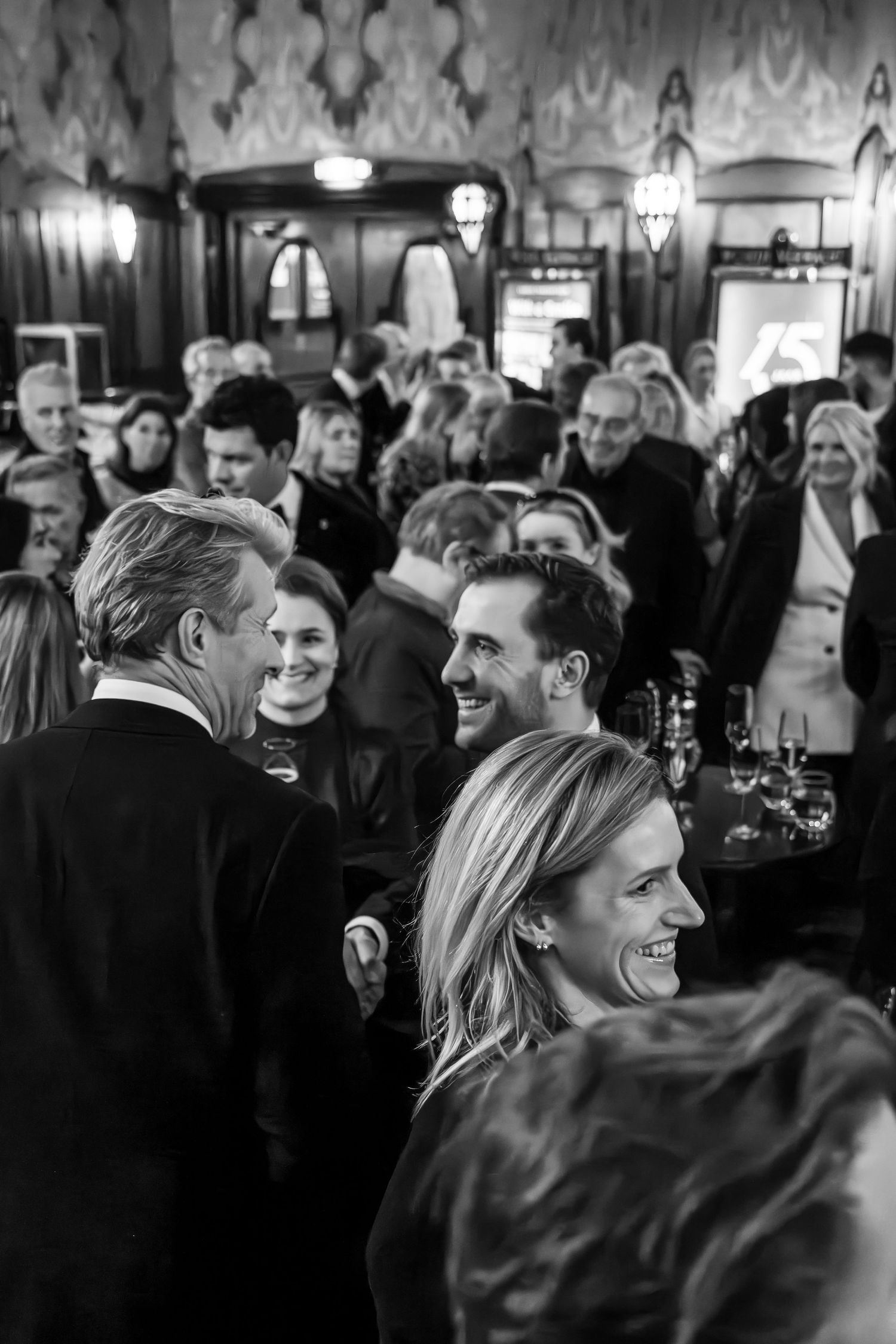 A black and white candid photo shows people laughing and socializing at an elegant evening event.