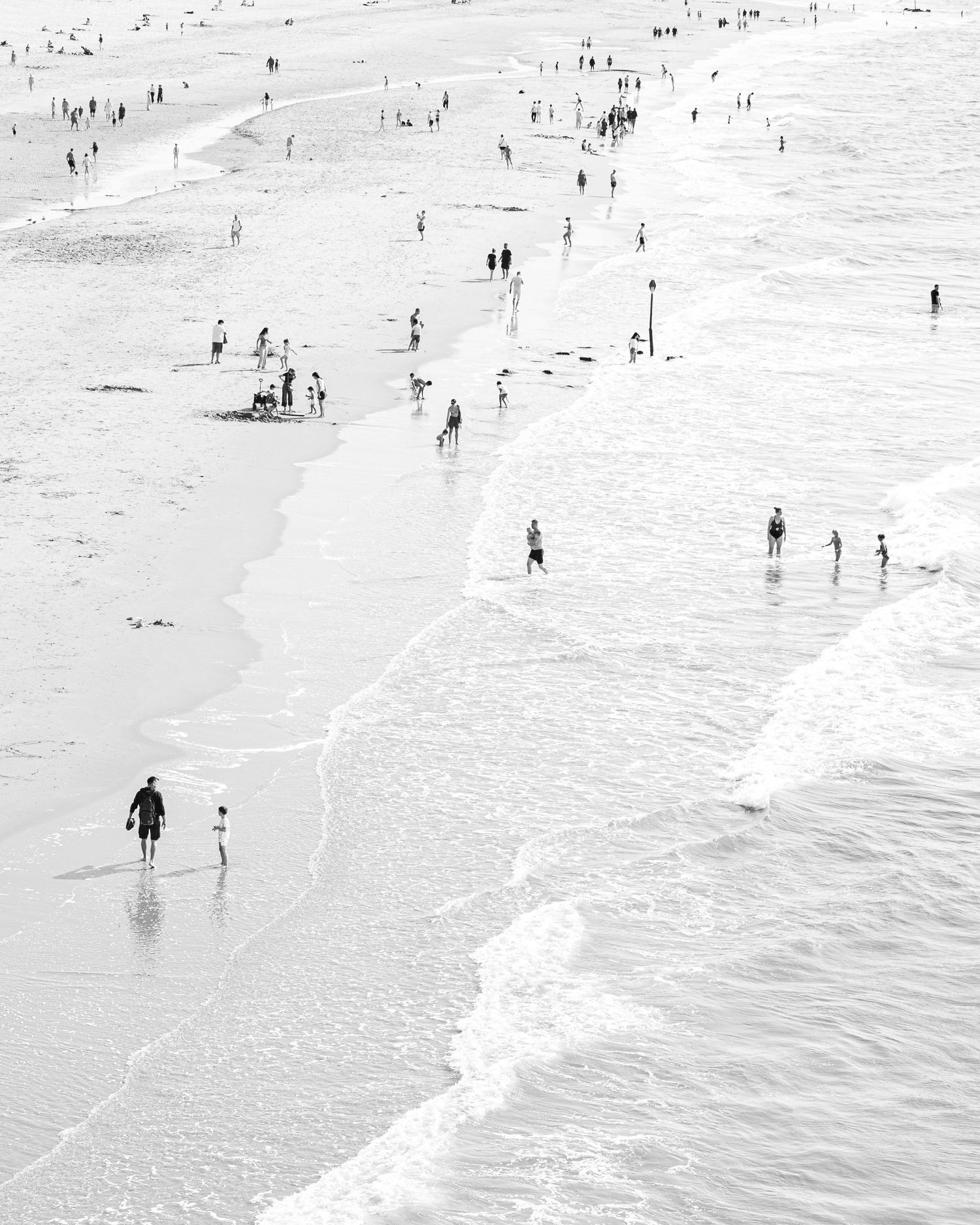 People enjoying a sunny day at a crowded beach with waves rolling onto the sand in a black and white photograph.