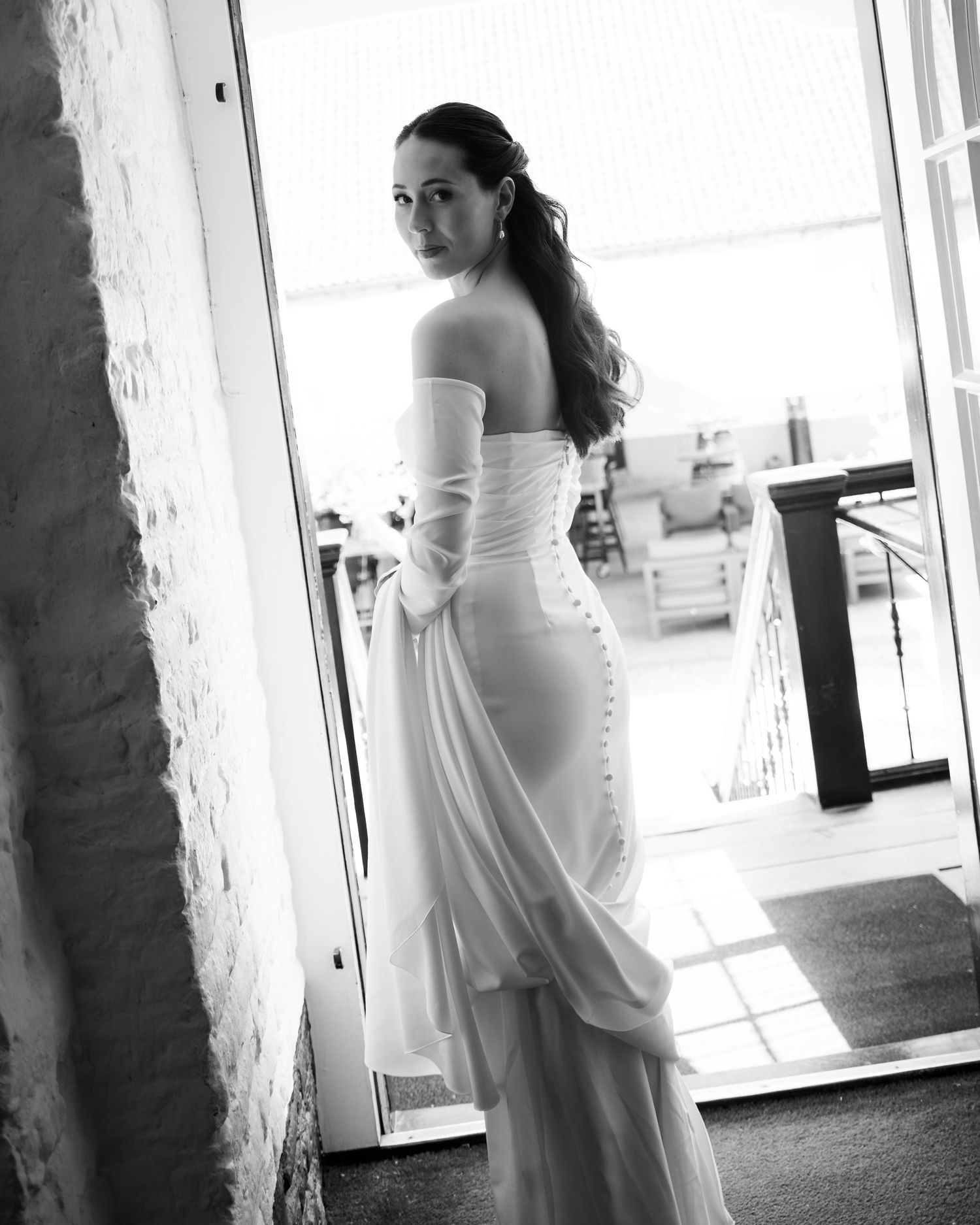 Black and white portrait of a bride in an elegant off-shoulder wedding gown looking over her shoulder in natural light.