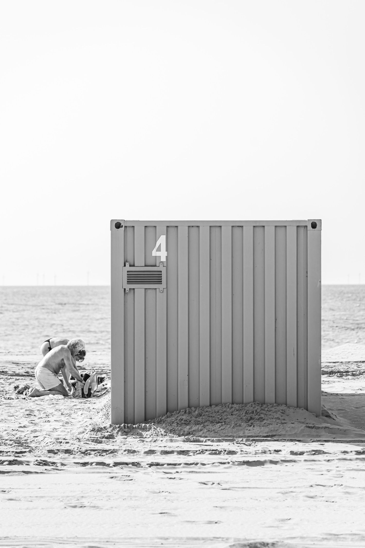 Black and white sequence showing someone gradually hiding behind a striped beach changing cabin on a sandy shore.