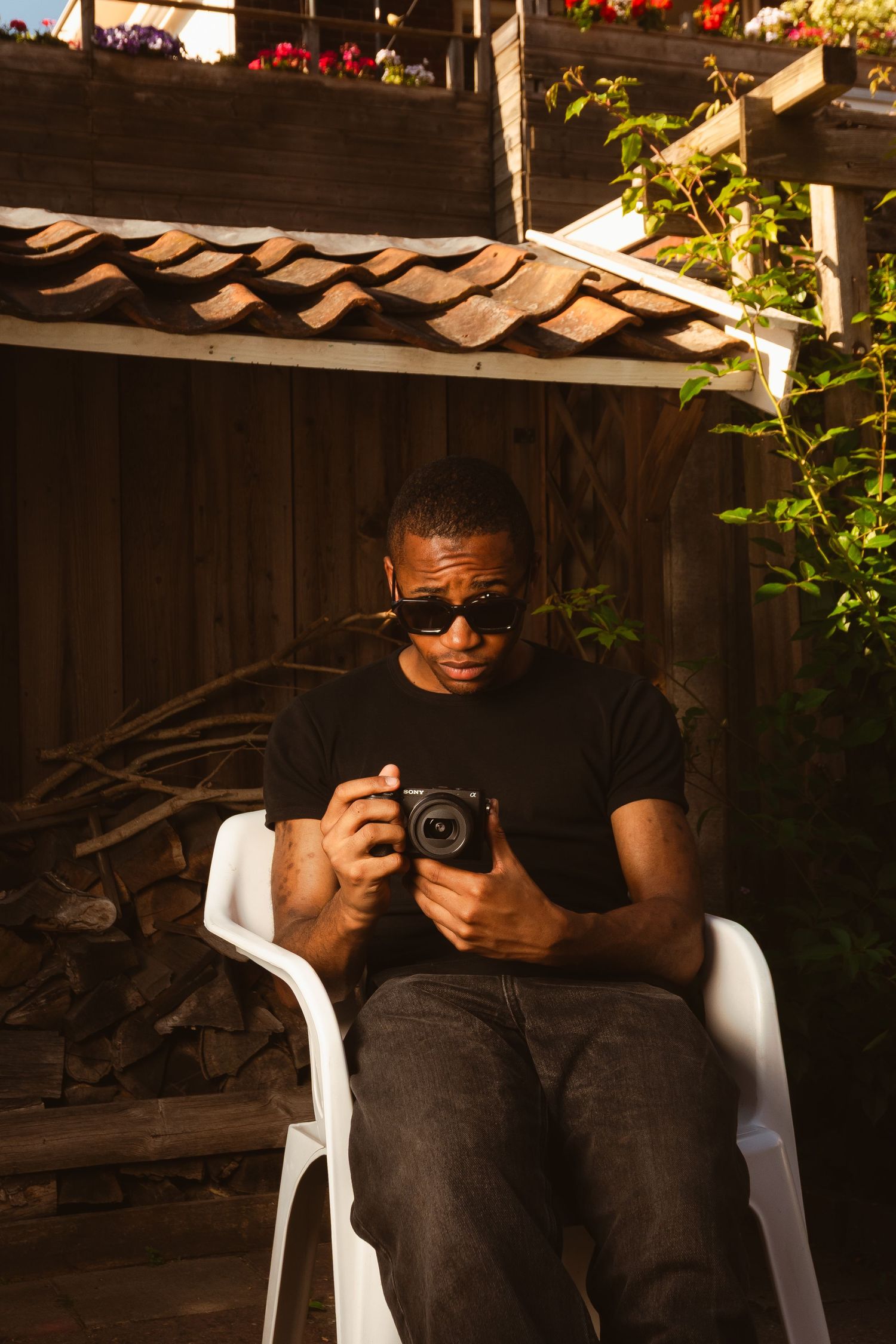 Person in sunglasses sits on white chair holding a camera in outdoor garden setting during golden hour lighting.