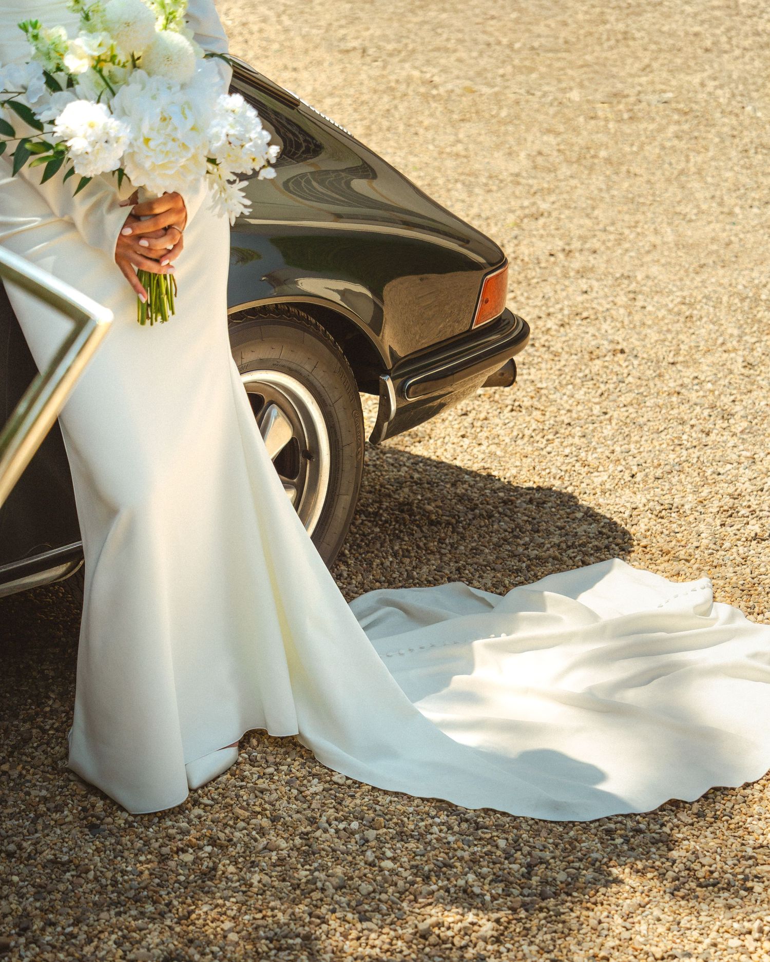 A flowing white wedding dress train and bouquet next to a classic black car on gravel ground.