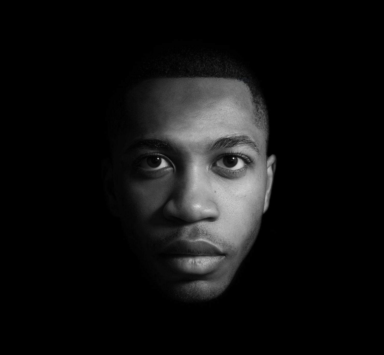 Dramatic black and white portrait of a young man's face emerging from darkness with intense gaze and sharp lighting.