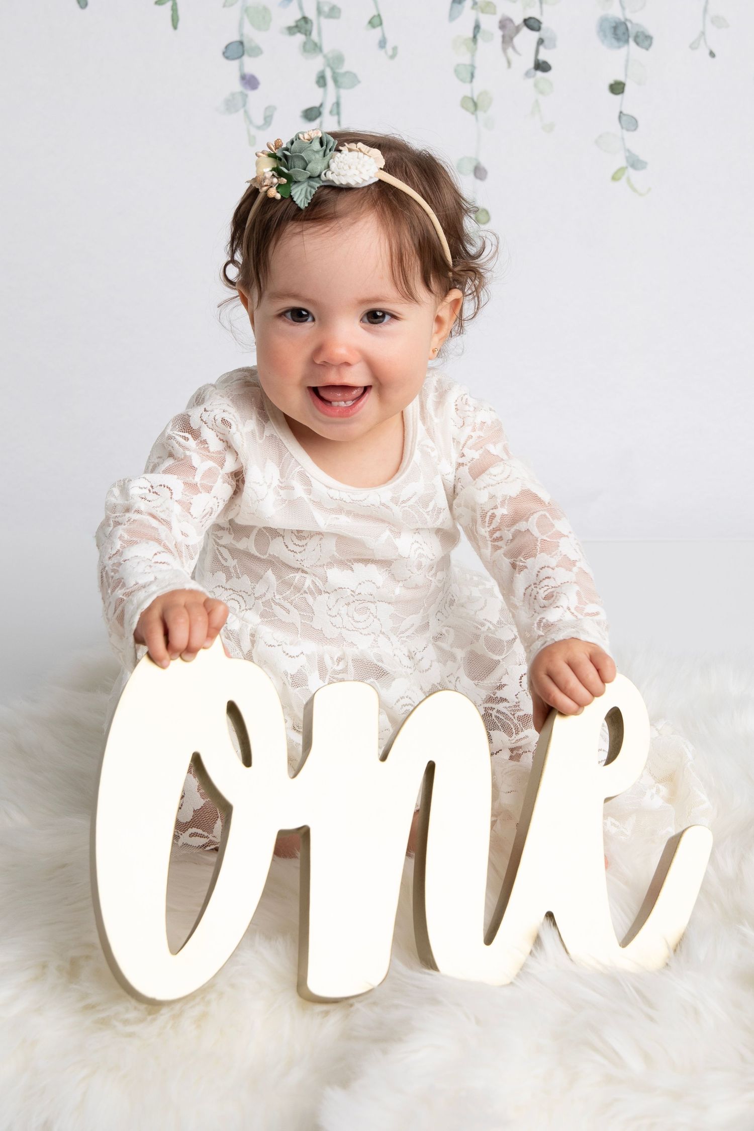 Baby in white lace dress sitting with decorative 'one' text letters against white background.