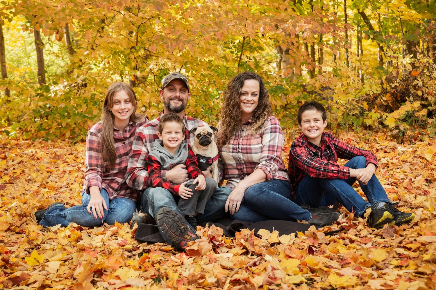 Family with dog sitting together on fallen autumn leaves wearing coordinated plaid outfits.