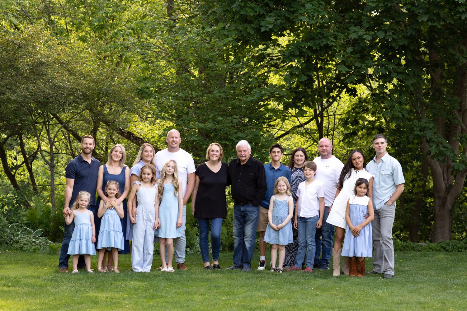 Large multigenerational family portrait outdoors on green lawn surrounded by lush trees in warm natural light.