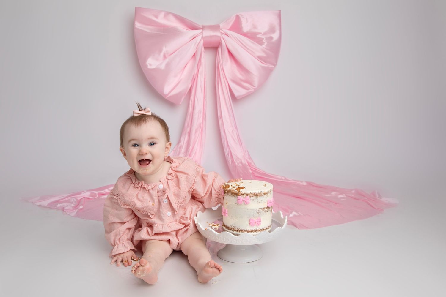 Baby in pink dress sits on floor with birthday cake and large pink bow backdrop for first birthday photoshoot.
