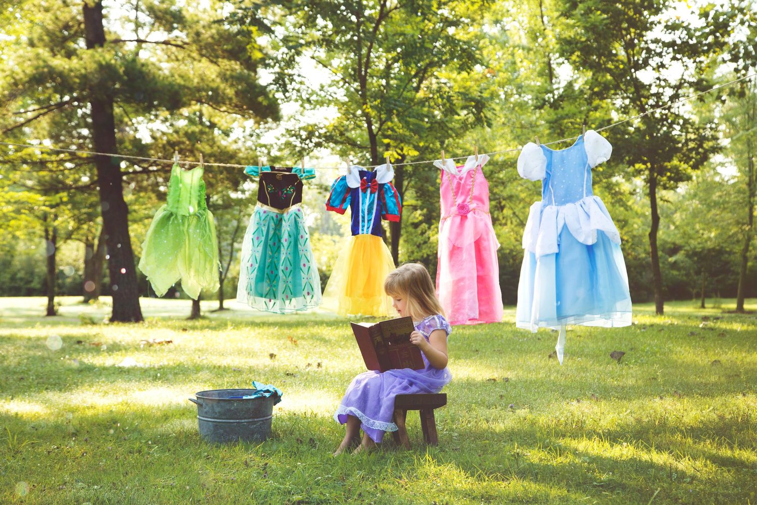 Colorful princess dresses hanging on clothesline outdoors between trees with basket and figure on bench below.