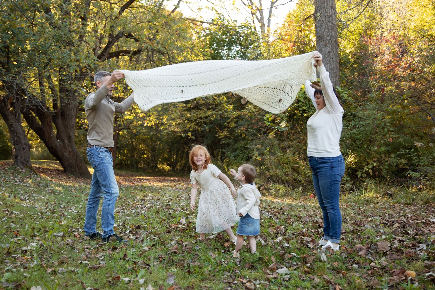 Parents and children playing under a white sheet in a wooded area during fall.