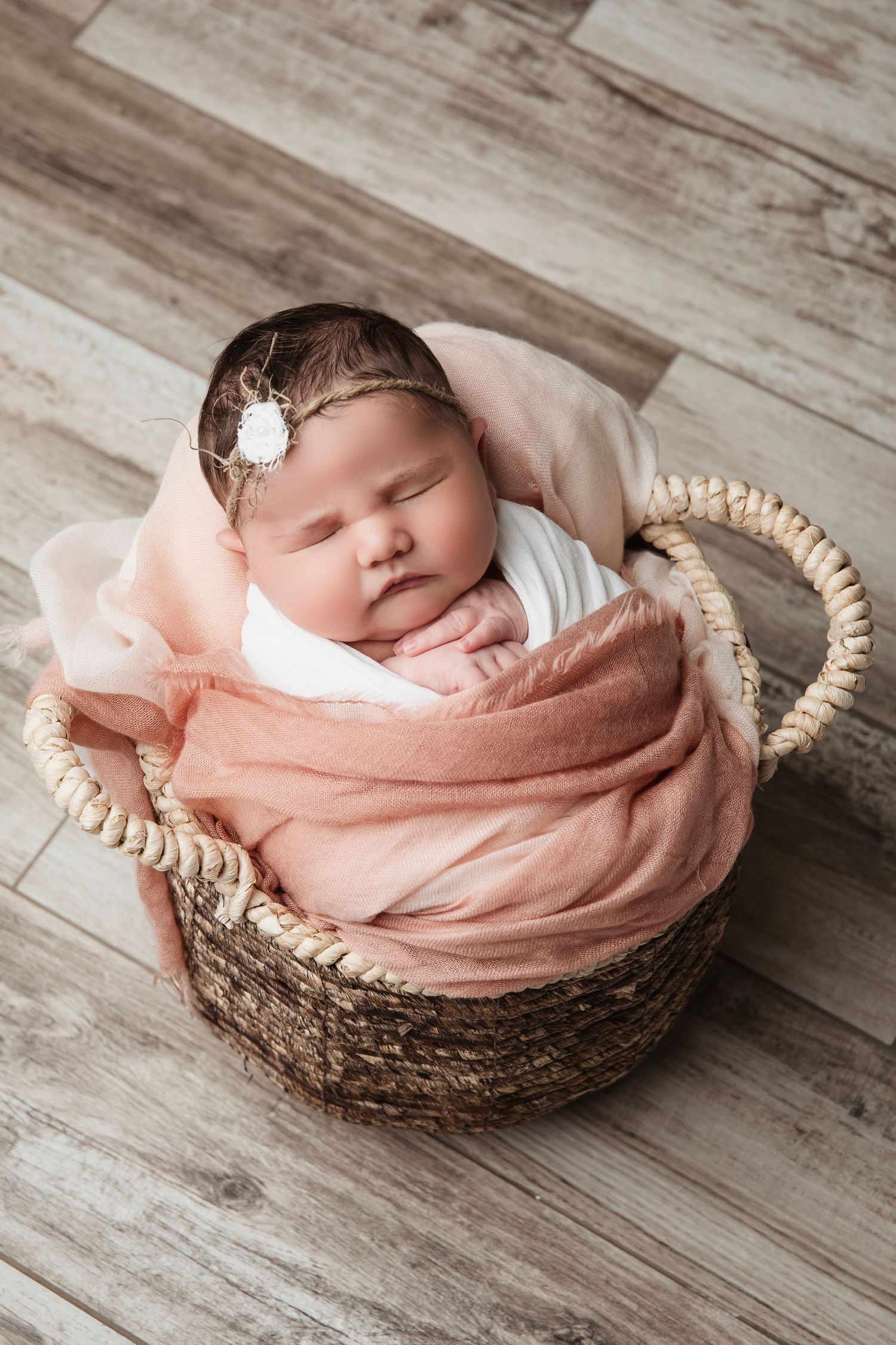 Peaceful newborn sleeping in woven basket with peach-colored wrap on wooden floor.