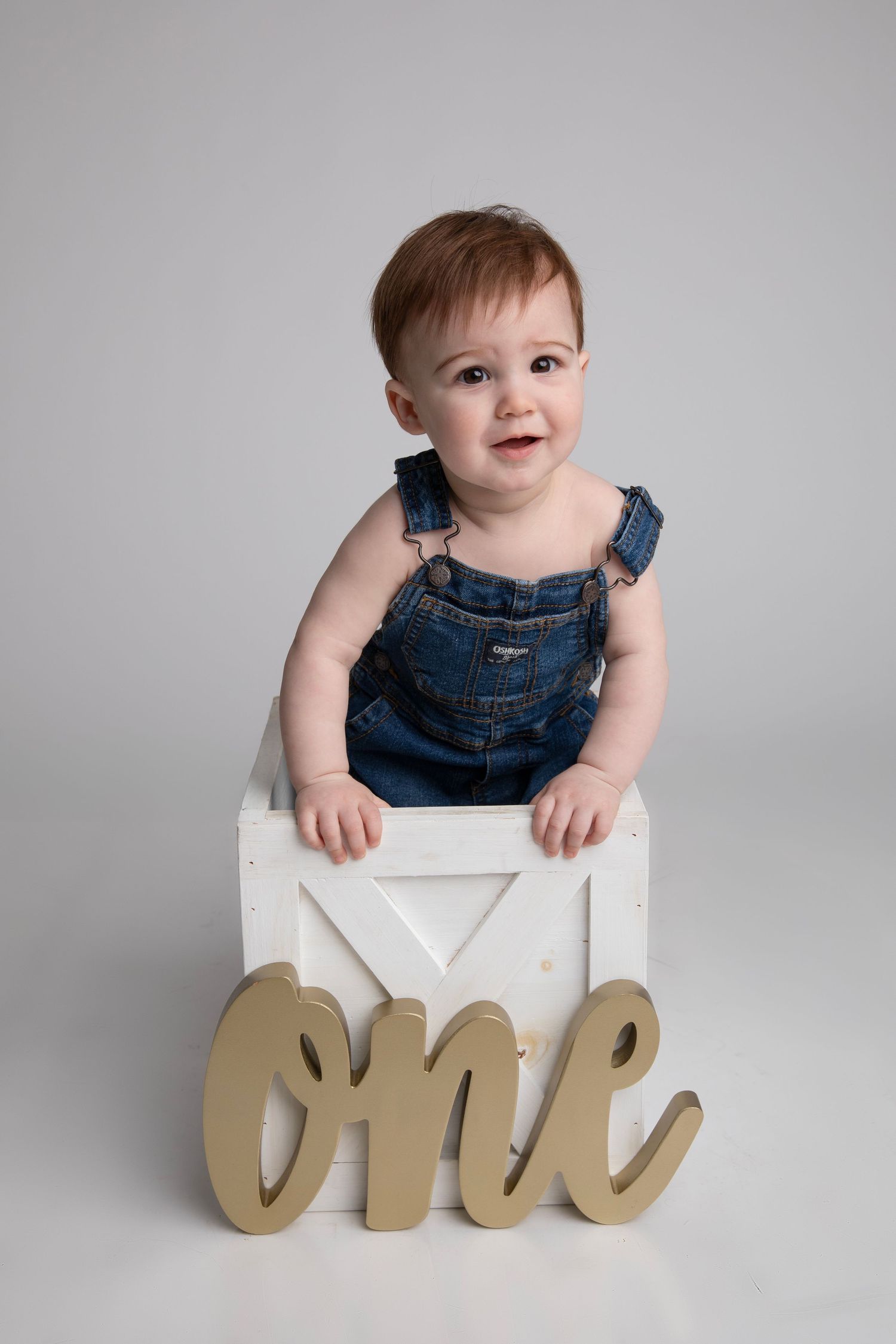 Happy baby in denim overalls sits on white box with gold 'one' sign against grey background.
