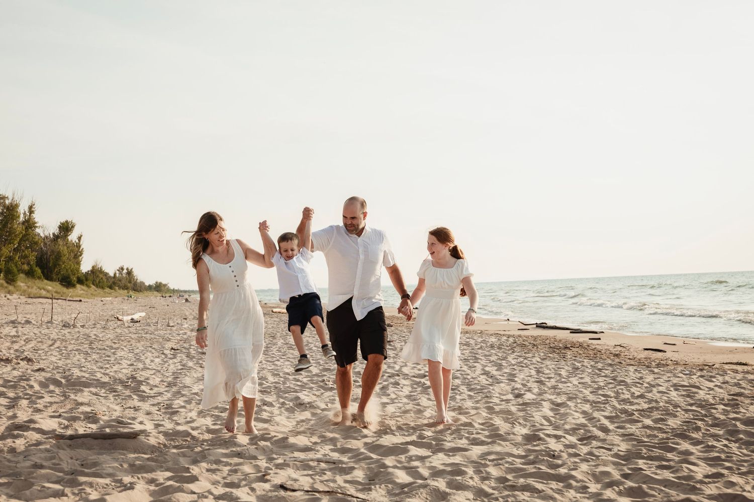 Family group in light colored clothing holding hands while joyfully walking together on a sunny beach.