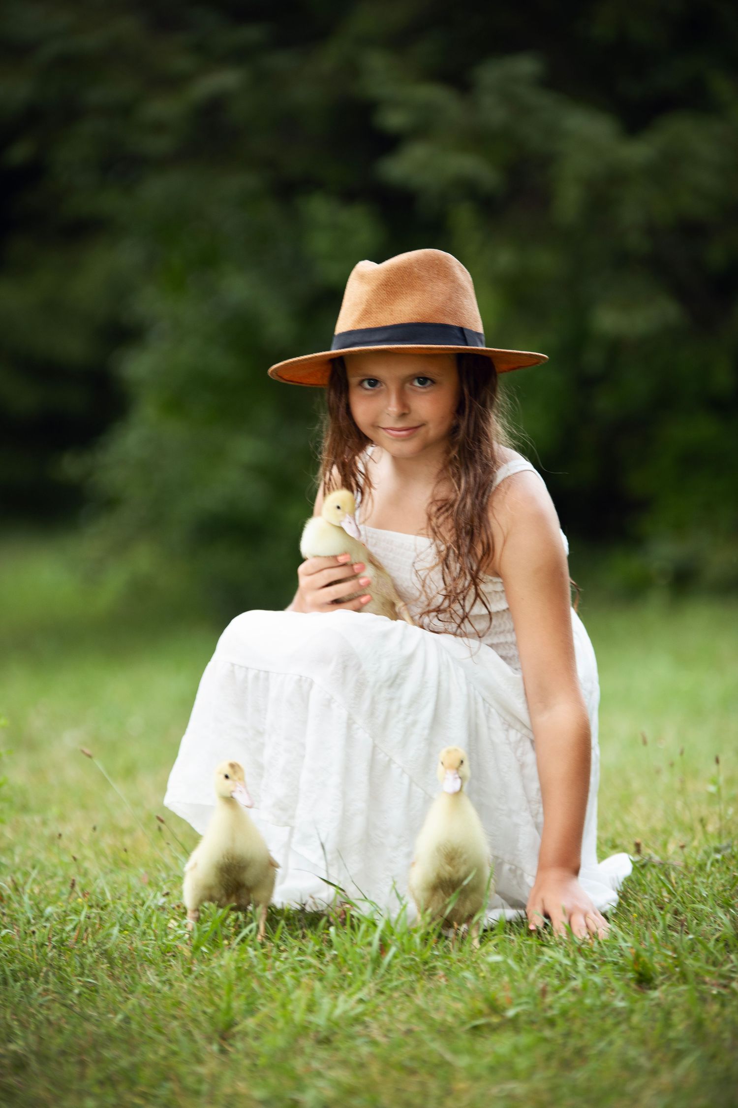 A small child wearing a white dress and brown wide-brimmed hat sits dreamily in a lush green meadow on a sunny day.