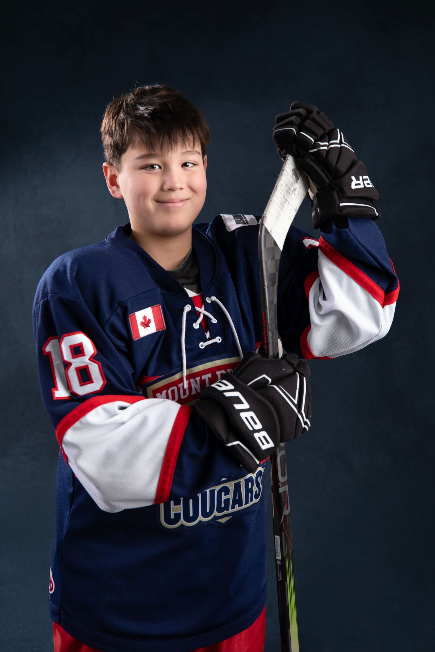 Young hockey player in navy Cougars jersey number 18 with Canadian flag patch, holding a Bauer stick, posing for team photo.