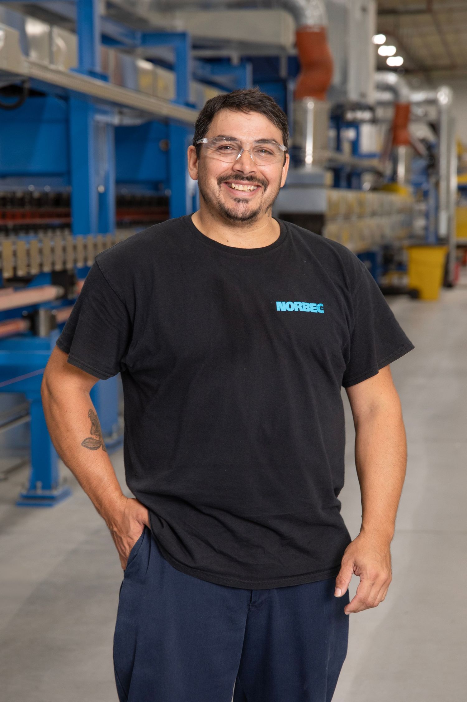 Warehouse worker in black shirt and navy pants standing in front of industrial storage shelves.