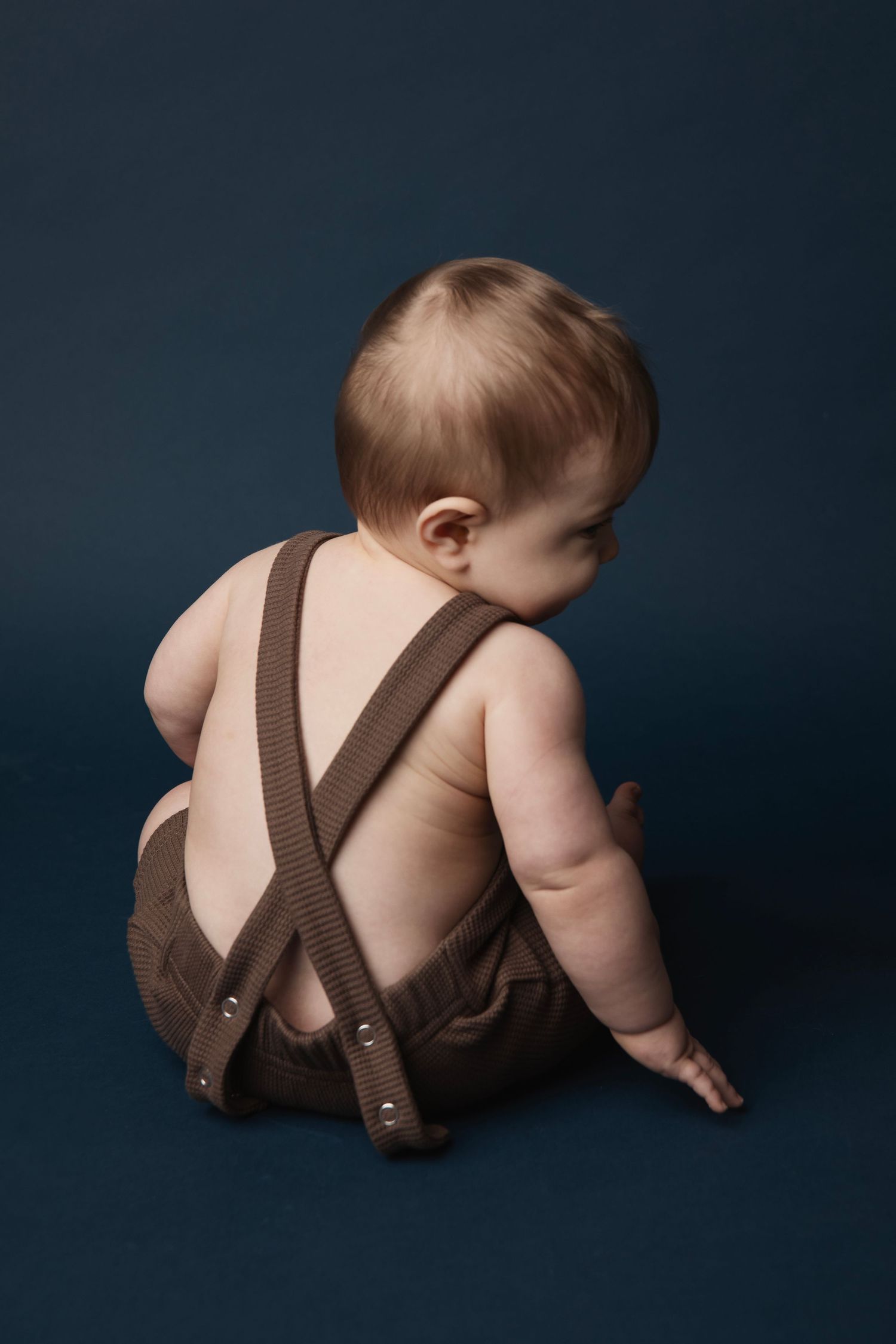 Small baby wearing brown overalls photographed from behind against dark backdrop in studio portrait.