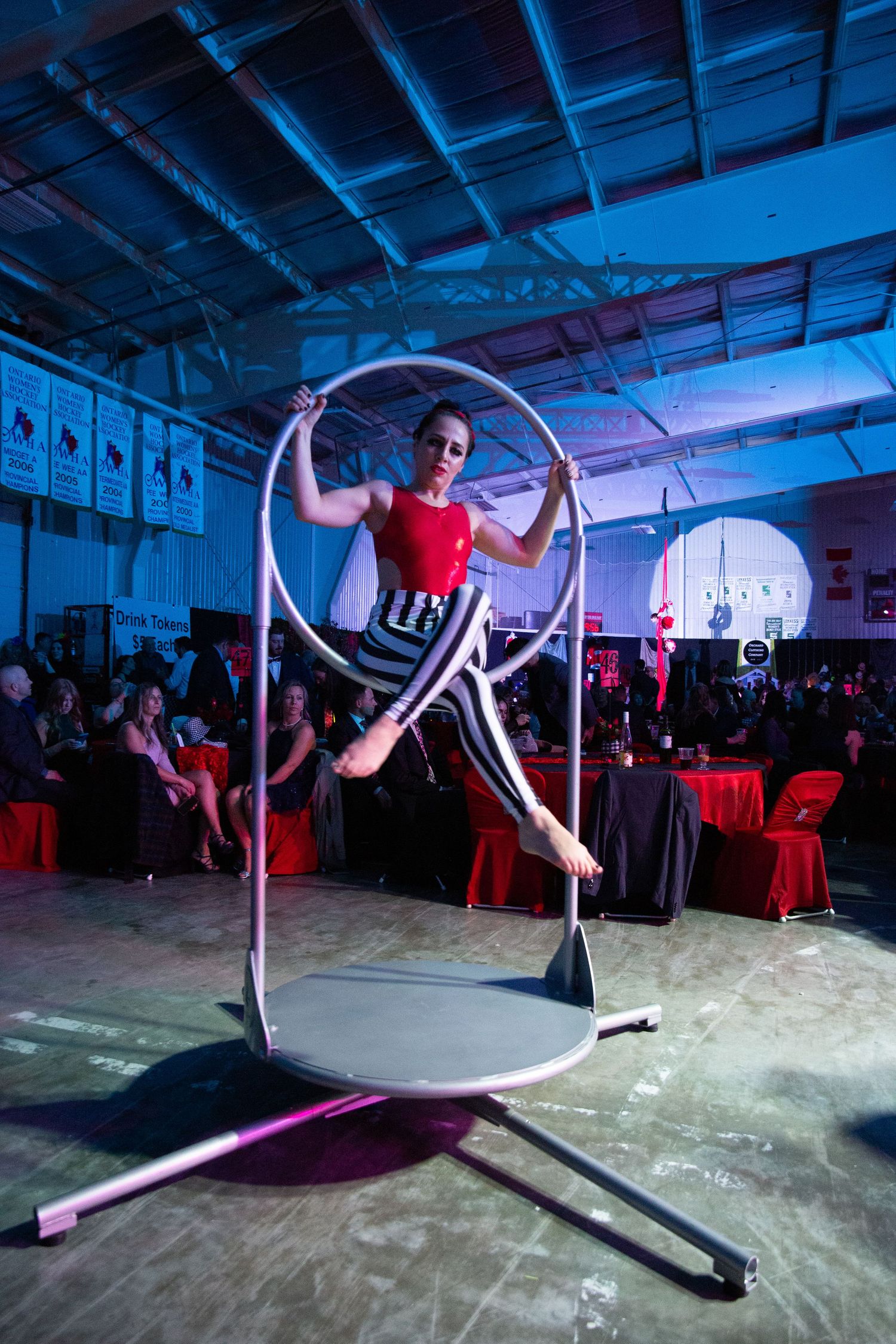 An aerialist performs a graceful pose on a freestanding circular apparatus in a venue with red-decorated tables.