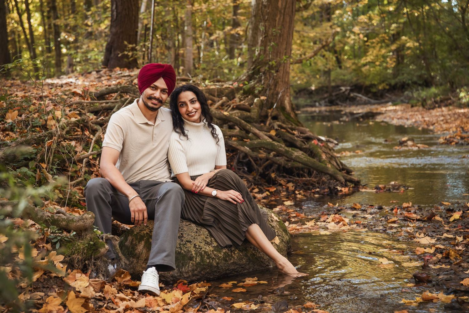A couple poses together near a creek surrounded by fallen autumn leaves in a forest setting.