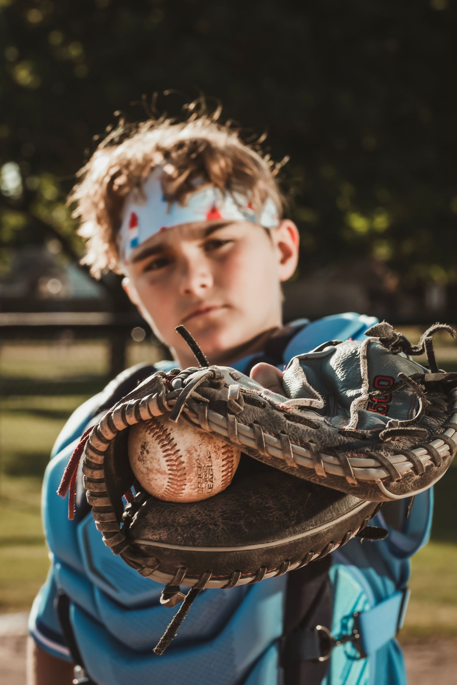 Young baseball player in blue uniform holds worn catcher's mitt with baseball toward camera outdoors.