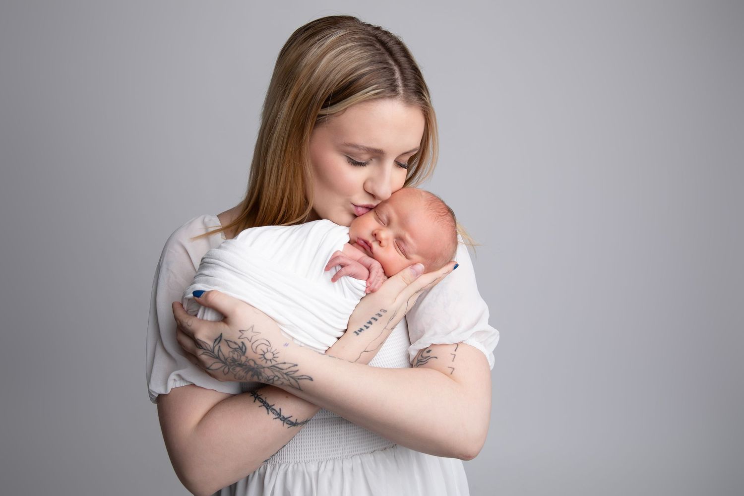 Tender moment as mother in white dress cradles and kisses sleeping newborn baby against grey backdrop.