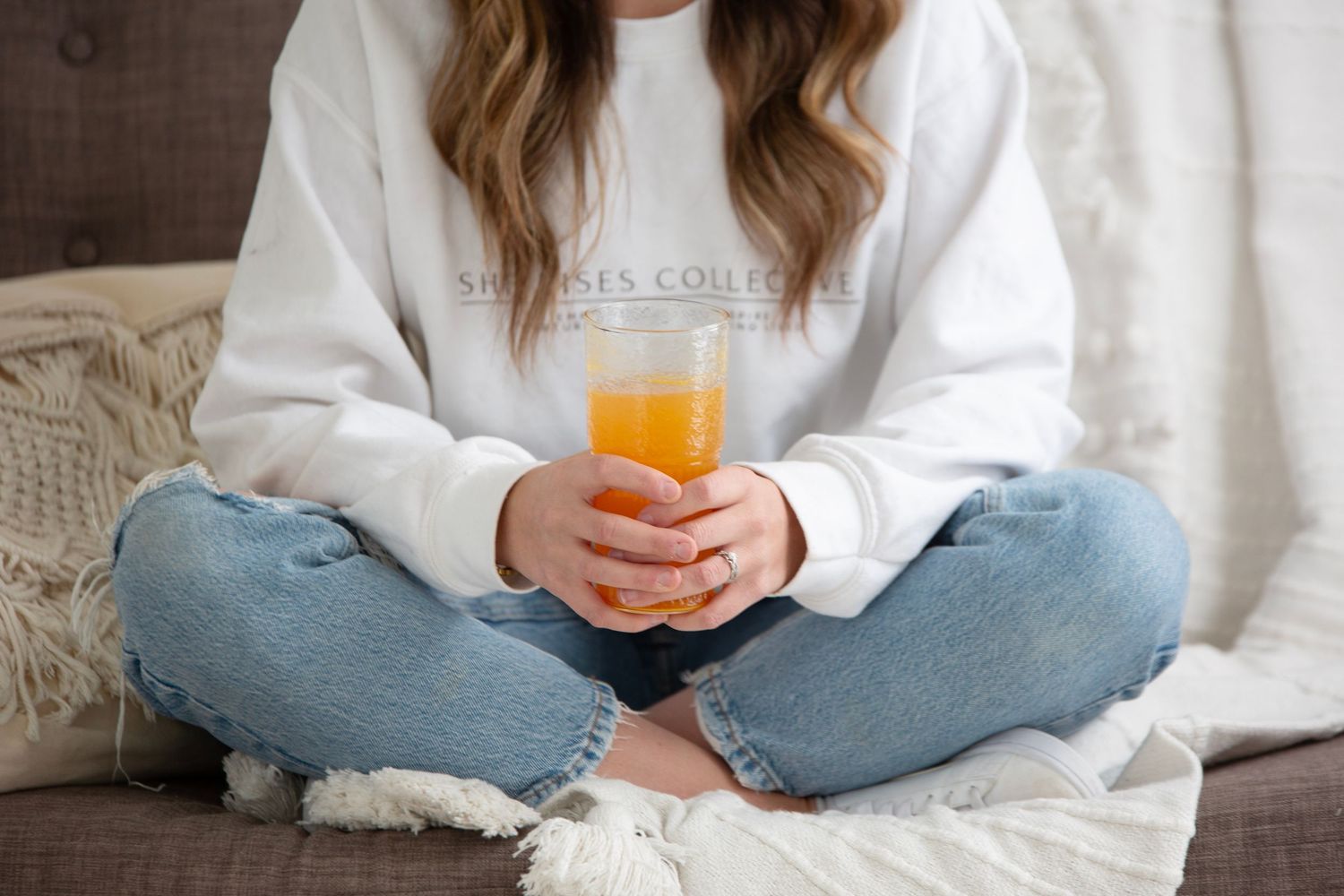 Person in white sweatshirt and jeans sitting cross-legged on couch holding glass of orange juice.