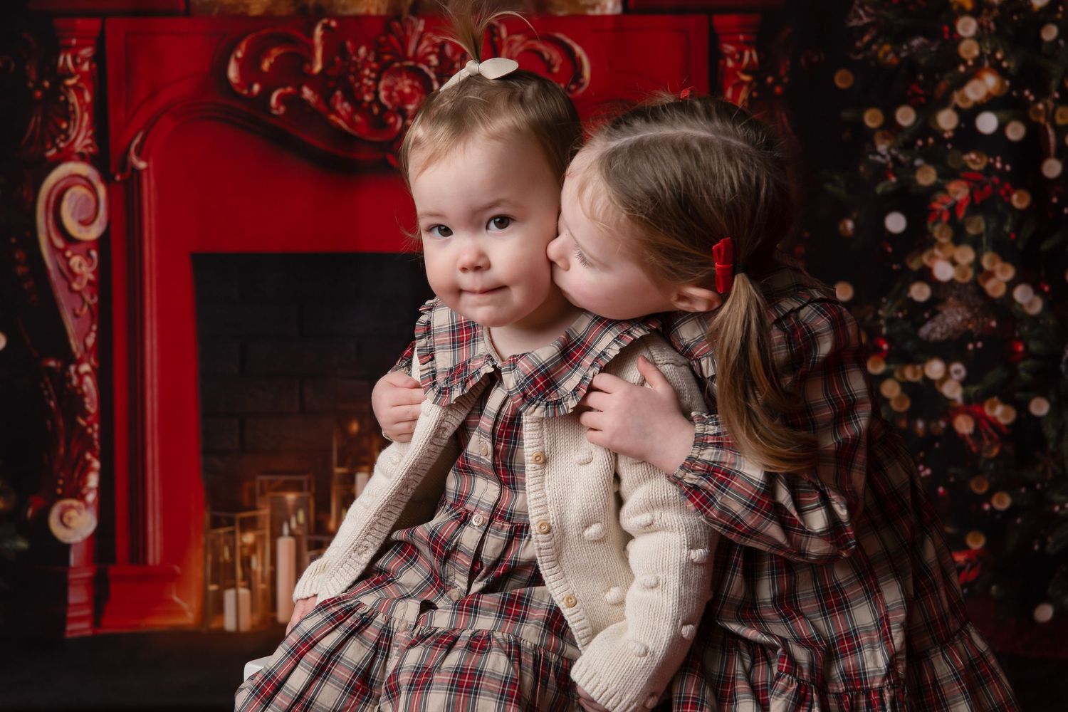 Two children in matching plaid outfits sit together by a red fireplace with holiday decorations in the background.