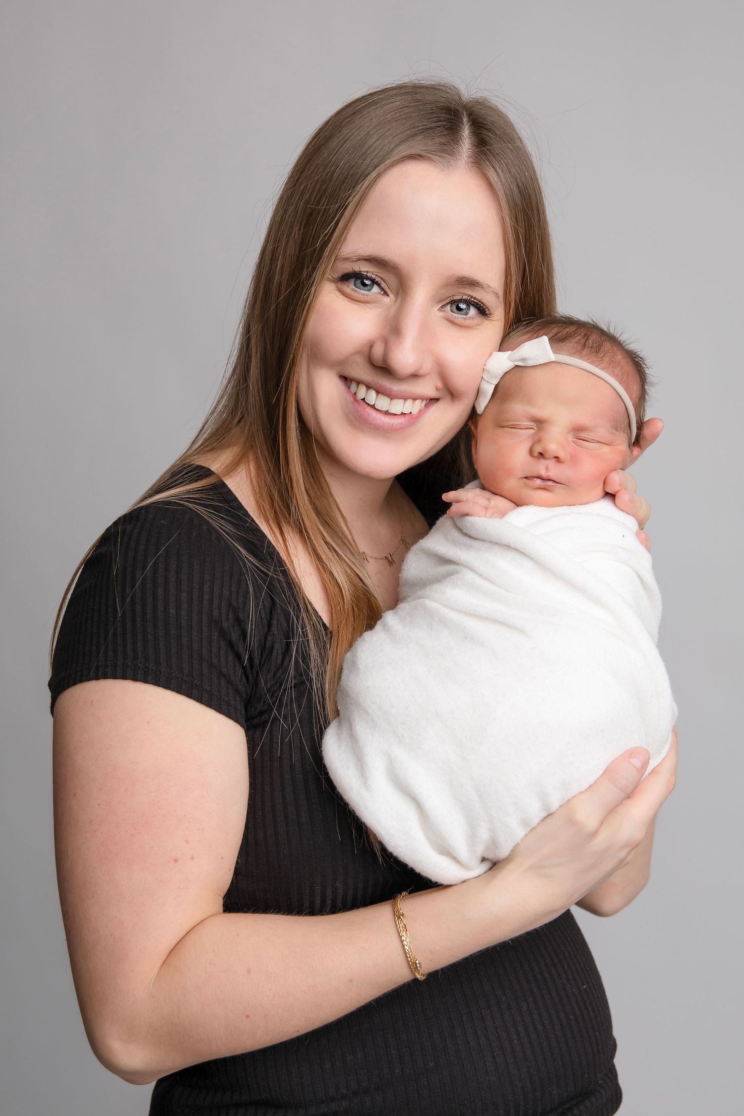 Mother in black shirt holding newborn wrapped in white blanket against gray backdrop.