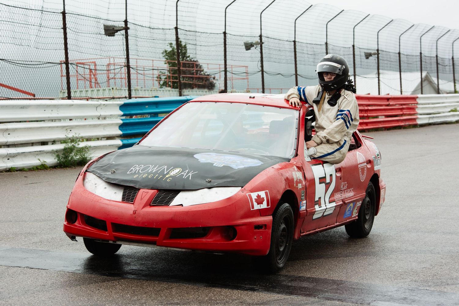 A red race car in the rain on a race track with blue and red safety barriers along the edge.