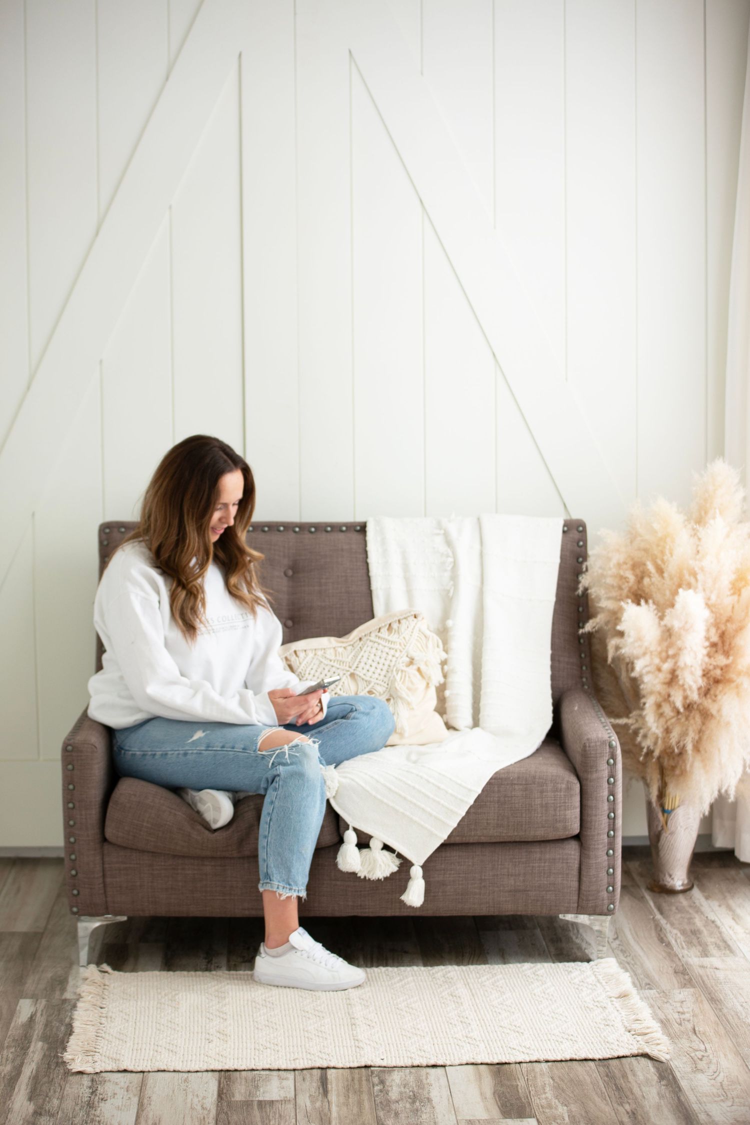 Woman in cozy white sweater and jeans sits on brown couch reading phone in minimalist room with pampas grass decor.