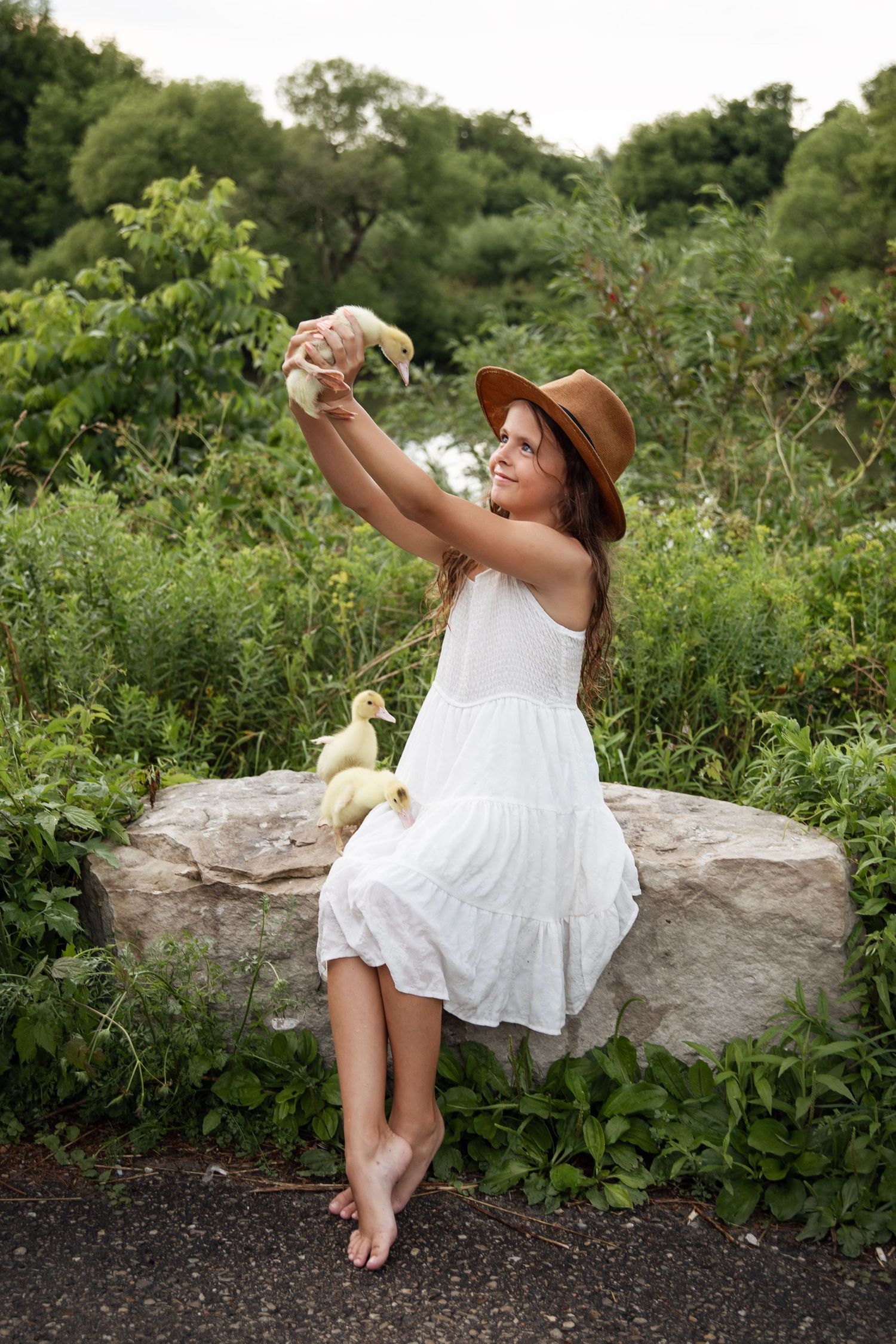 A person in a white summer dress and brown hat sits on a rock wall while taking a selfie in a lush garden setting.
