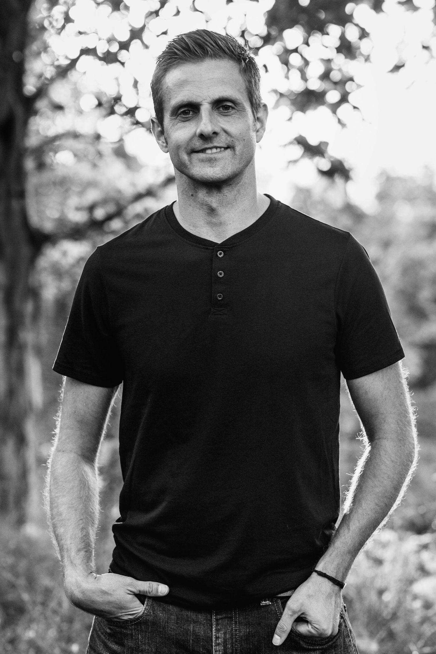 A black and white portrait of a person wearing a henley t-shirt and jeans in an outdoor setting with bokeh background.