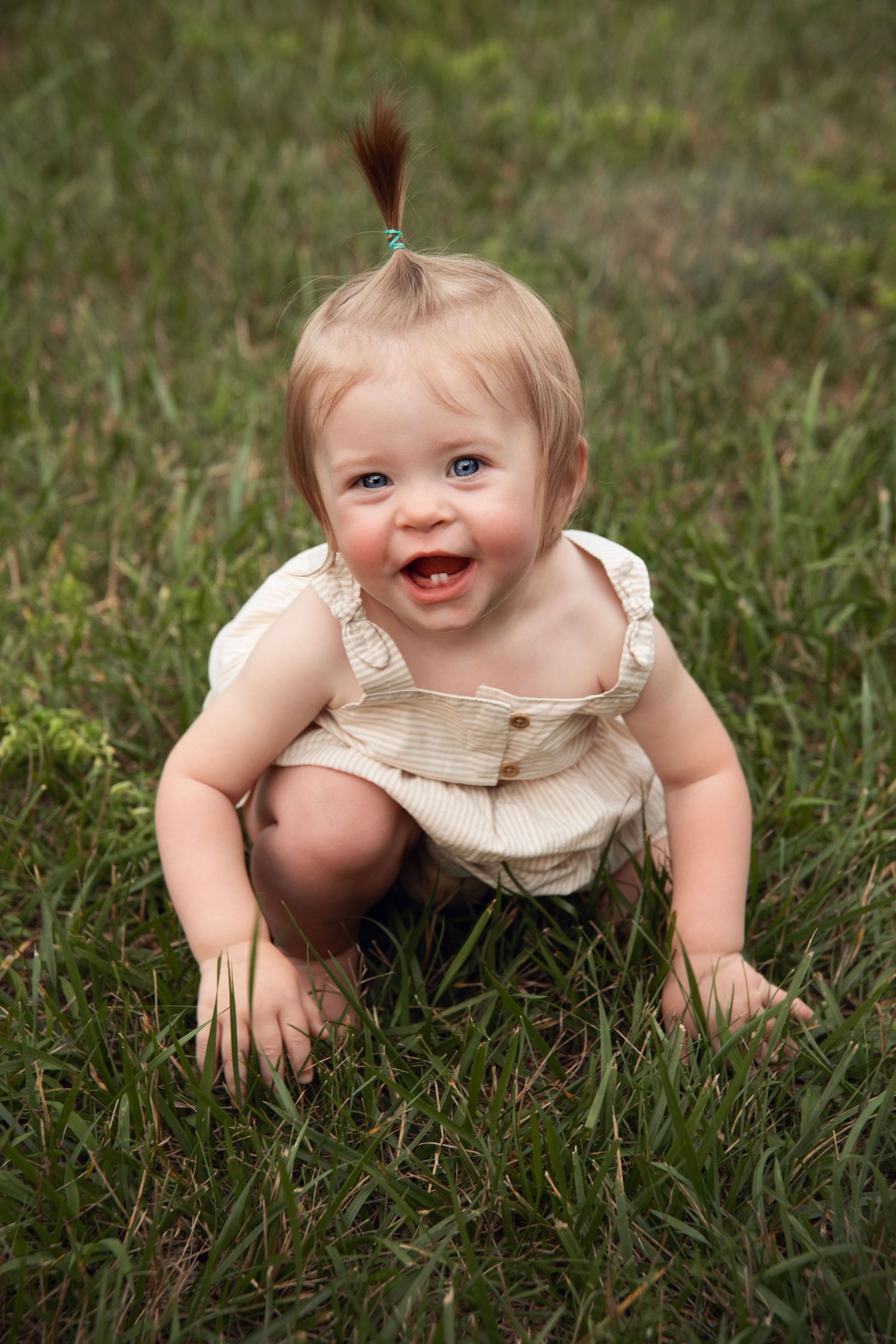 A laughing baby in a beige romper crouches in green grass with a tiny ponytail on top of their head.