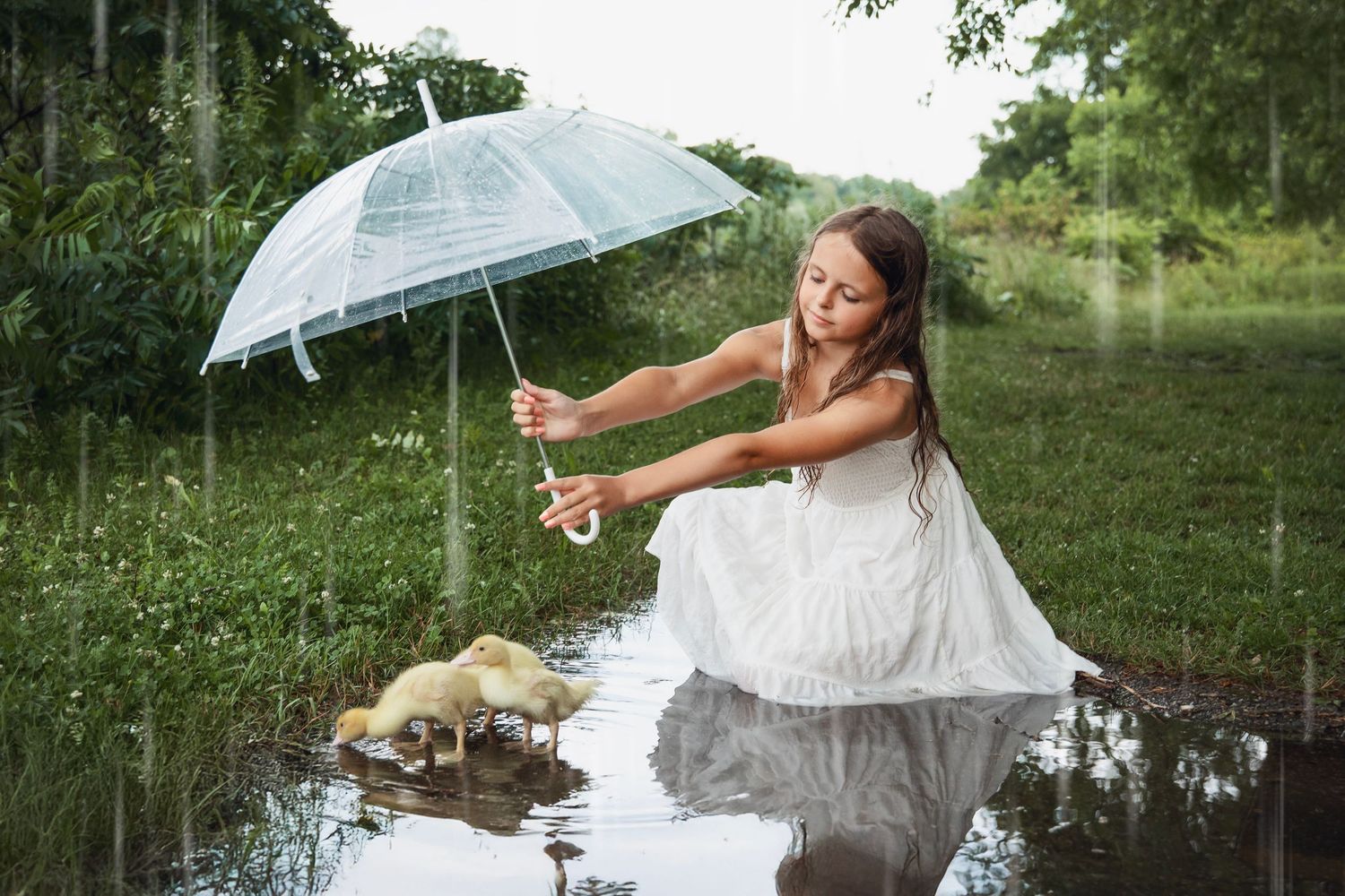 A person in a flowing white dress holds a clear umbrella while posing in front of a serene pond on a summer day.