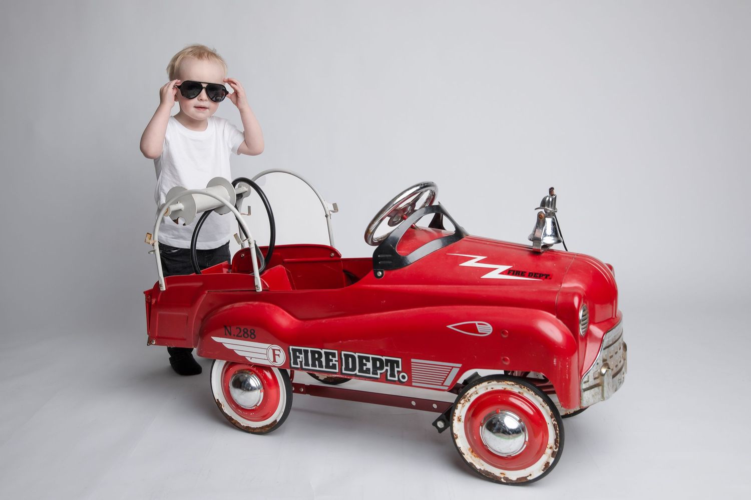 A child wearing sunglasses poses with a vintage red pedal car.