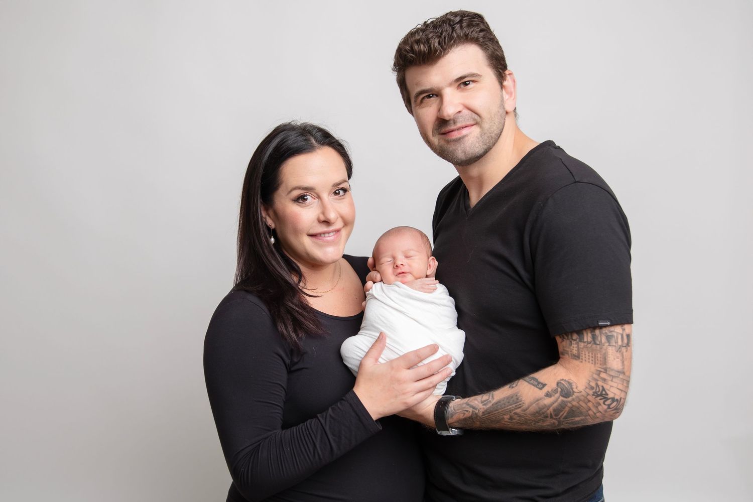 Family portrait featuring couple in black holding their newborn baby in white wrap.