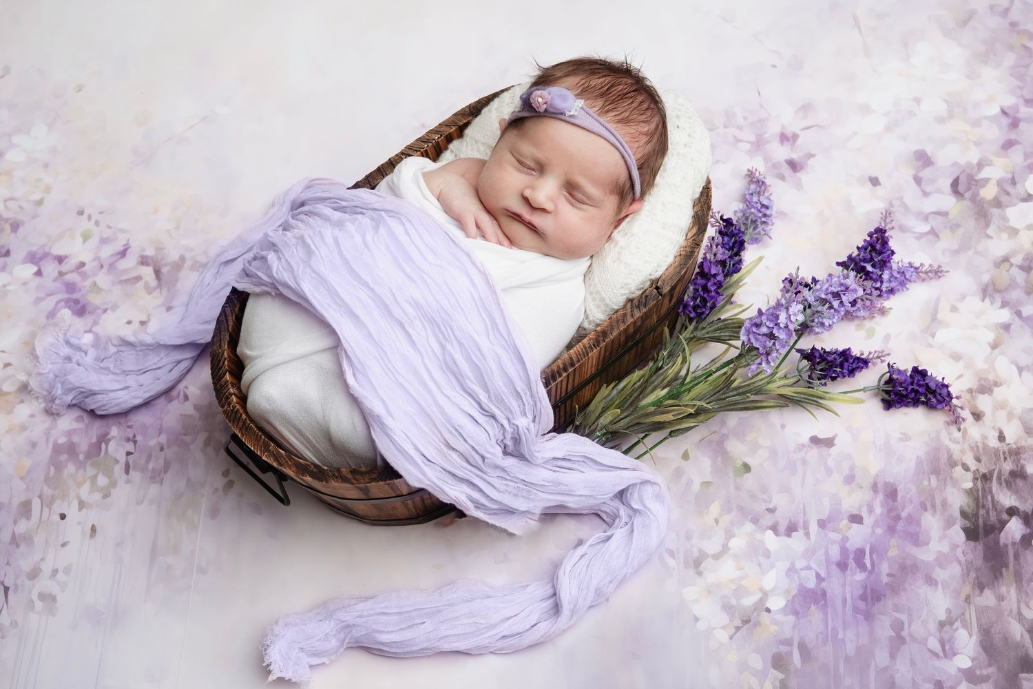 Peaceful newborn sleeping in woven basket with lavender sprigs and purple toned backdrop.