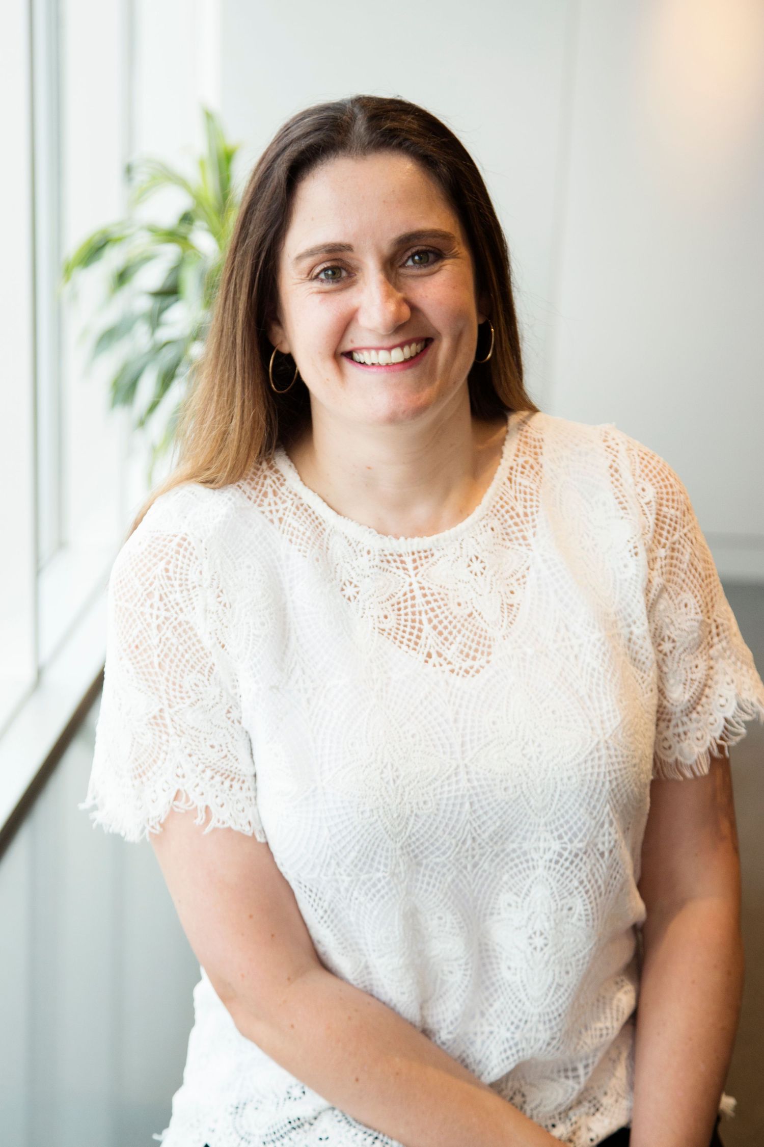 Professional headshot of someone wearing a white lace blouse standing near a window with a plant in the background.