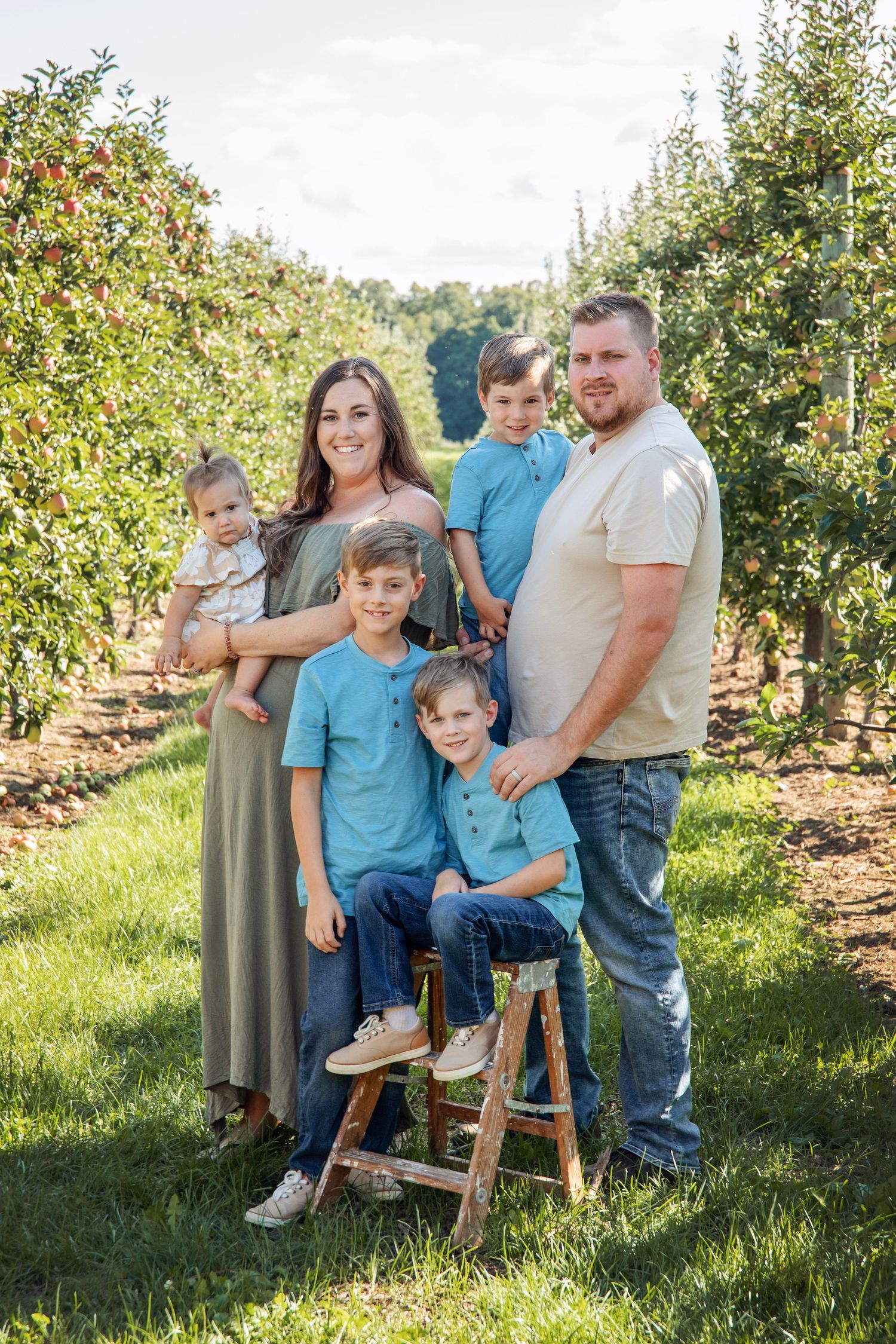 A family poses together on a rustic bench in an apple orchard during a sunny afternoon photo session.