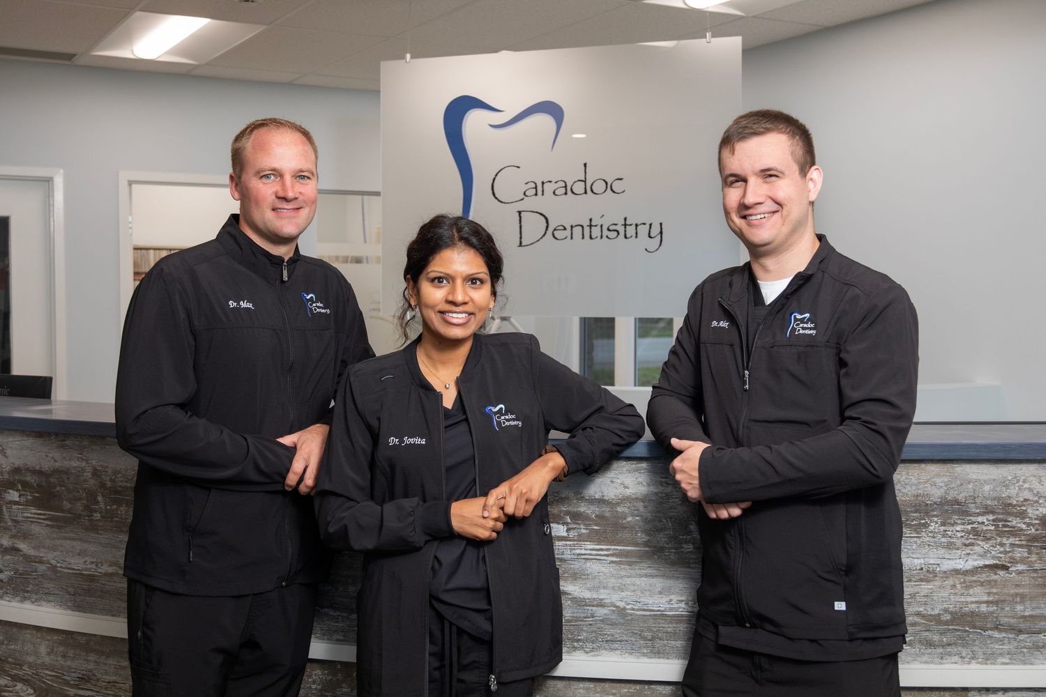 Three dental professionals in black scrubs stand in front of a Canalside Dentistry office sign and marble counter.