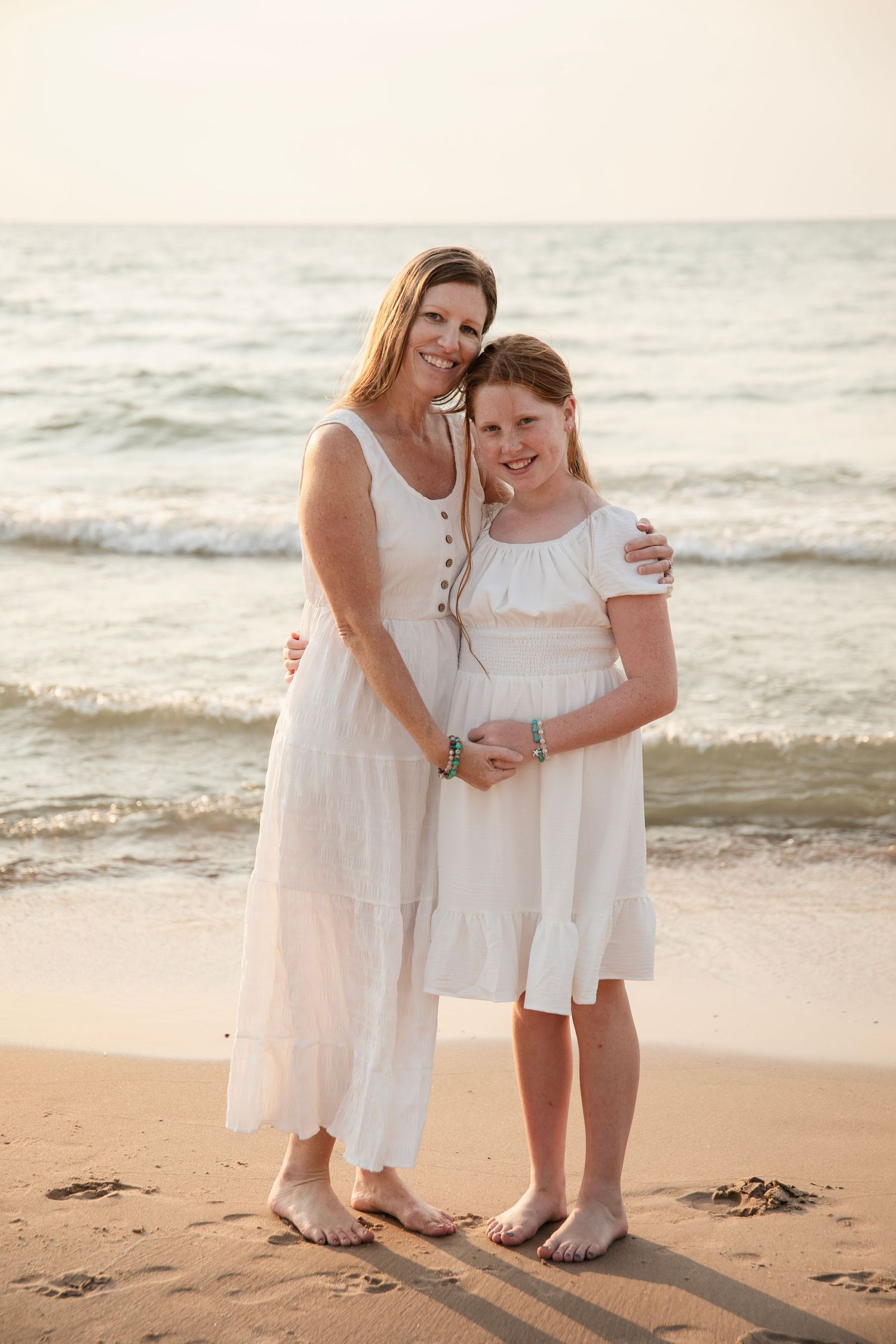 Two people wearing white dresses embrace on a sandy beach at sunset while waves roll in behind them.