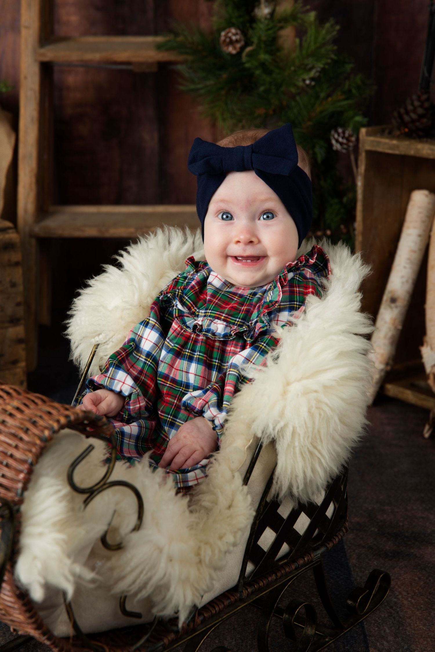 A smiling baby wearing plaid clothing sits in a cozy white fur-lined sleigh during a holiday photo session.