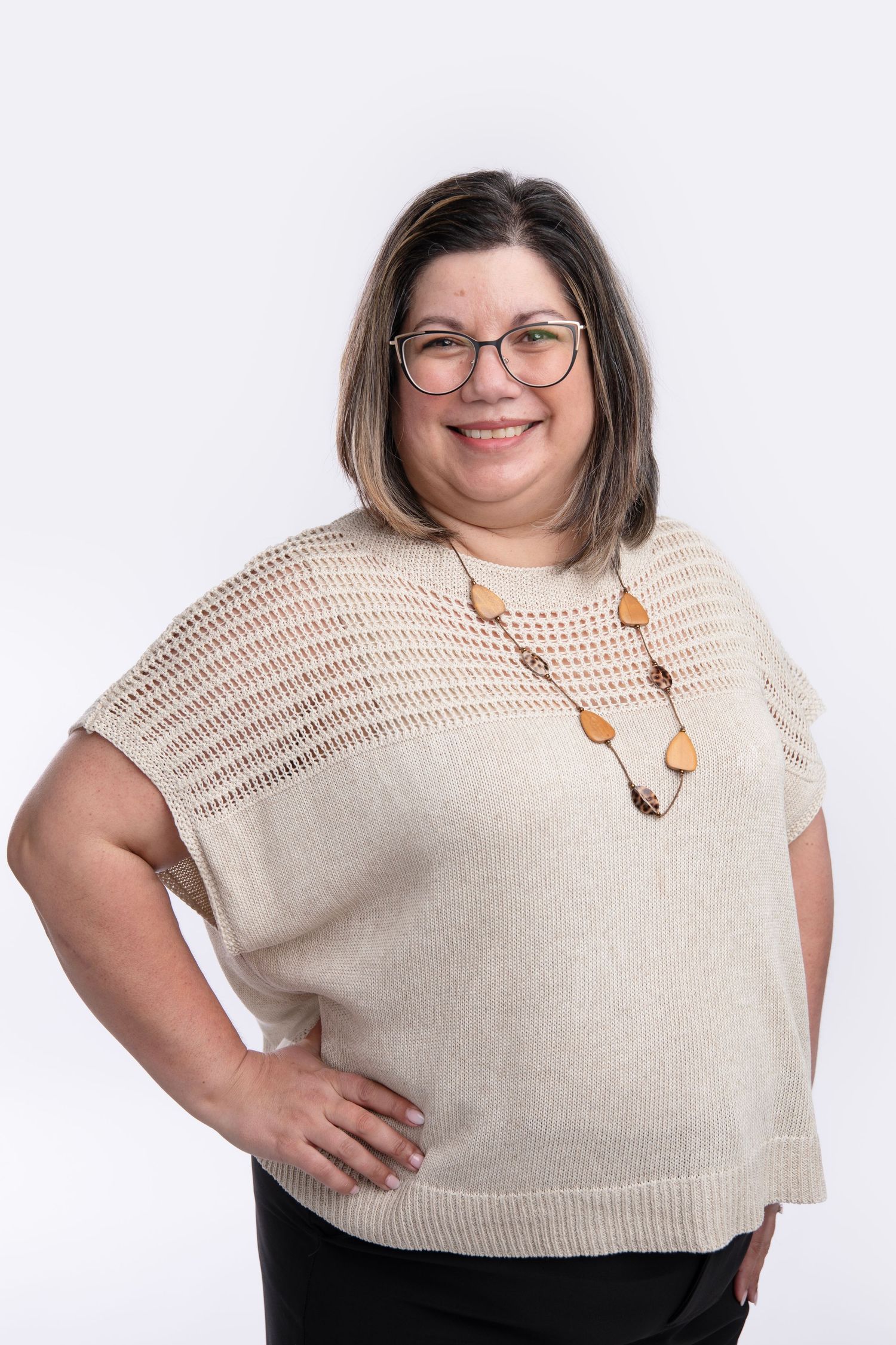 A smiling person wearing glasses and a cream knit sweater with wooden necklace posing against white background.