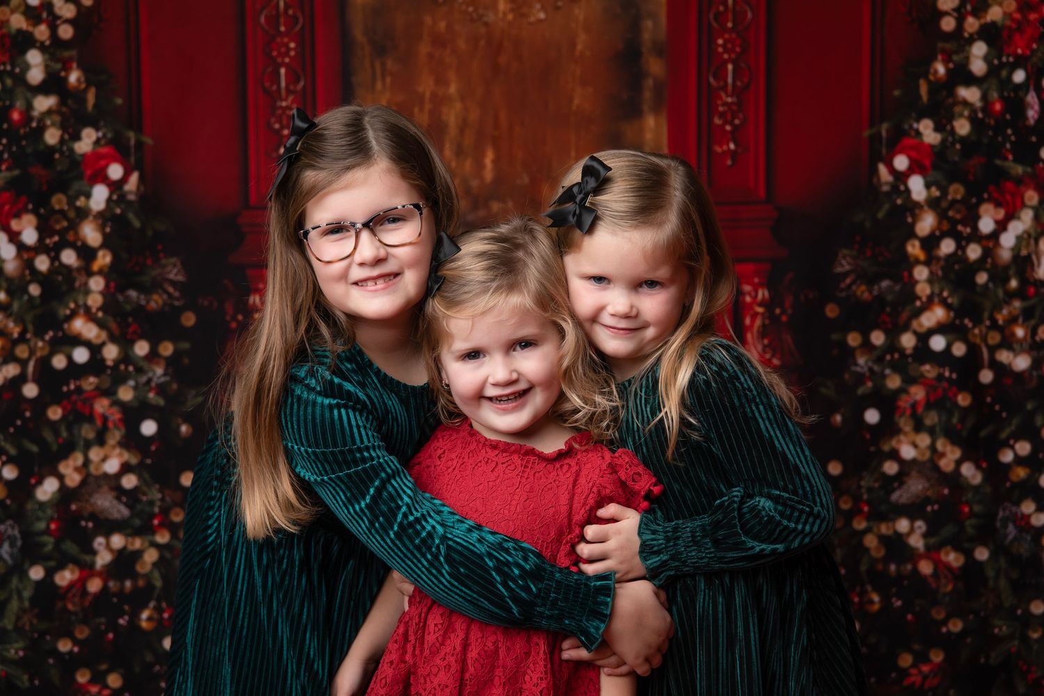 Three children in holiday attire pose together against a red backdrop with floral decorations.