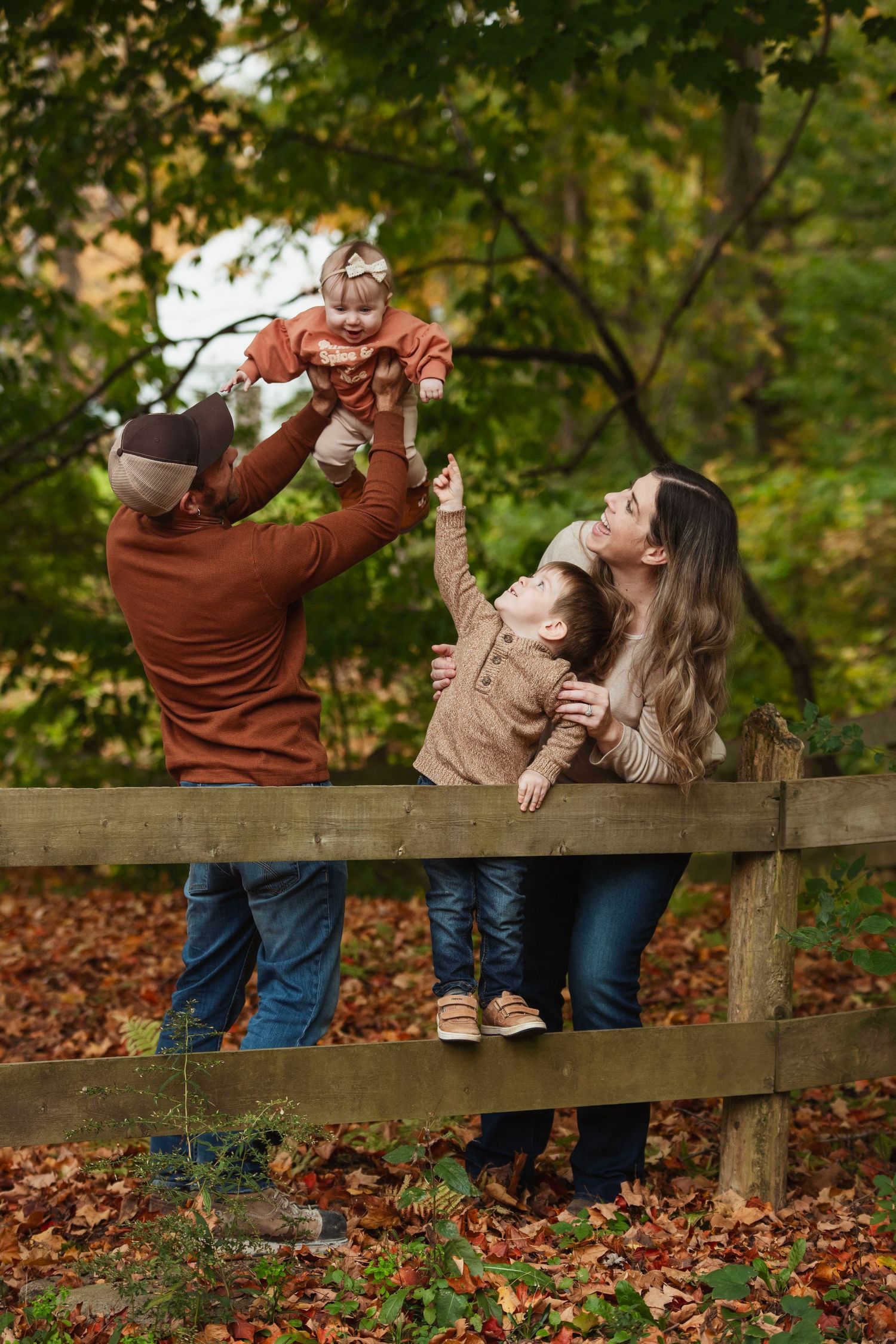 Family posing playfully along wooden fence in autumn setting wearing coordinated outfits.