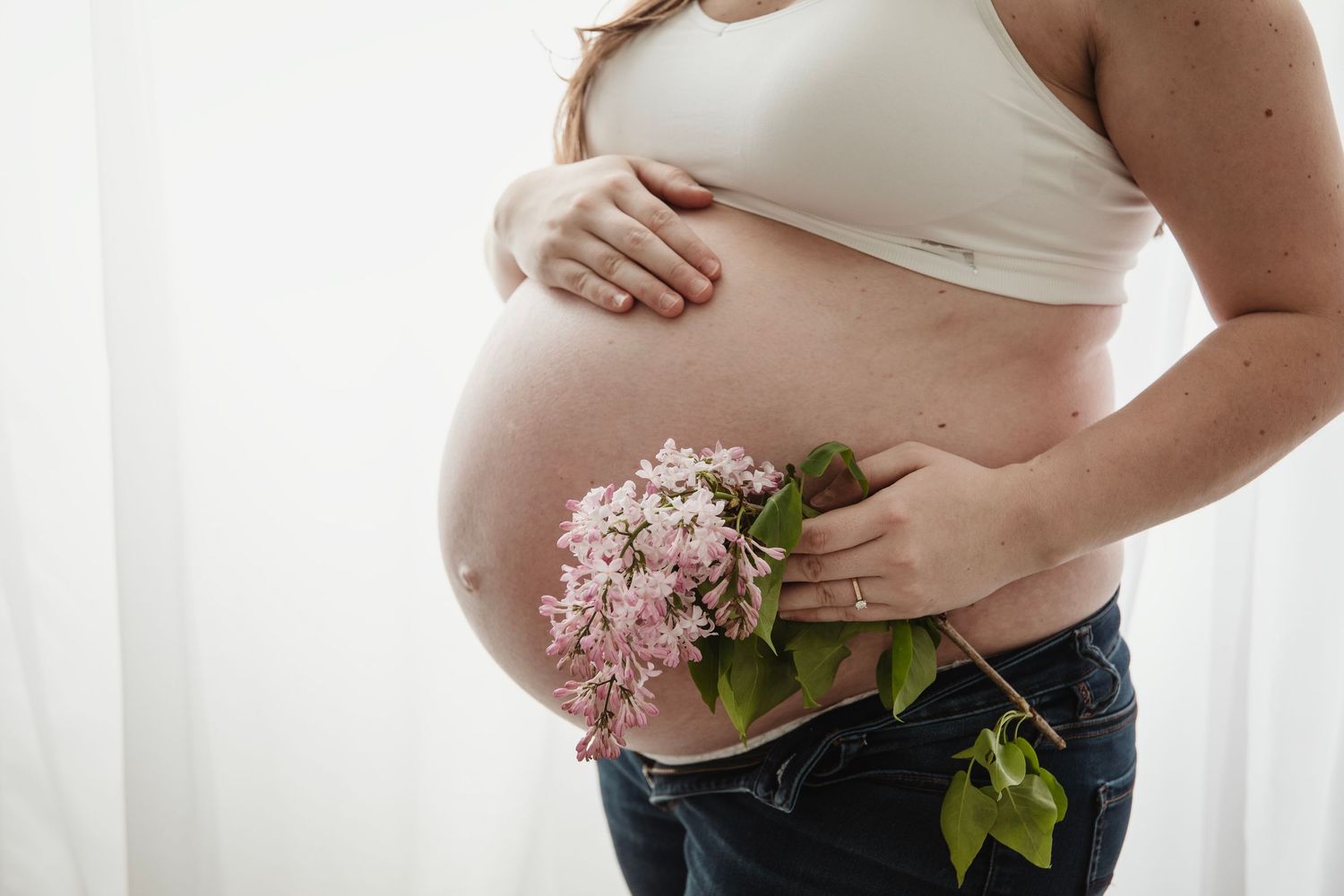 Side view of pregnant belly with pink flowers against white backdrop in intimate portrait.