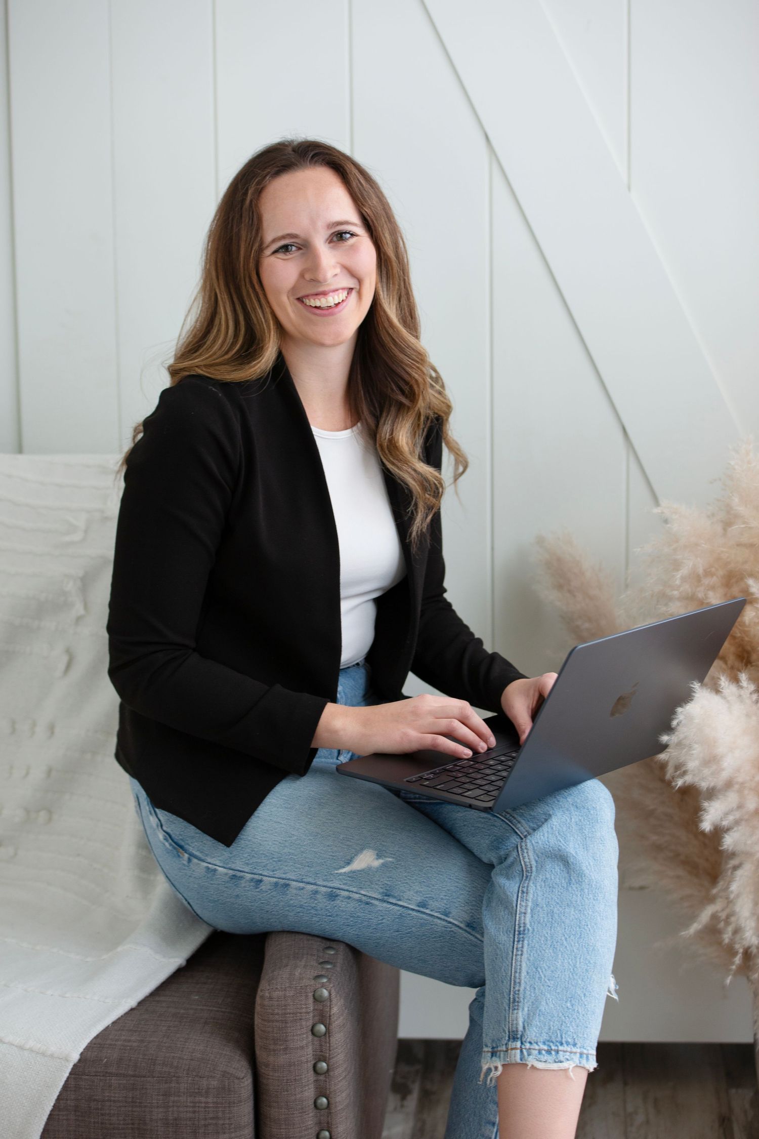 Professional wearing black blazer and jeans works on laptop while seated on ottoman against white wall with pampas grass decor.