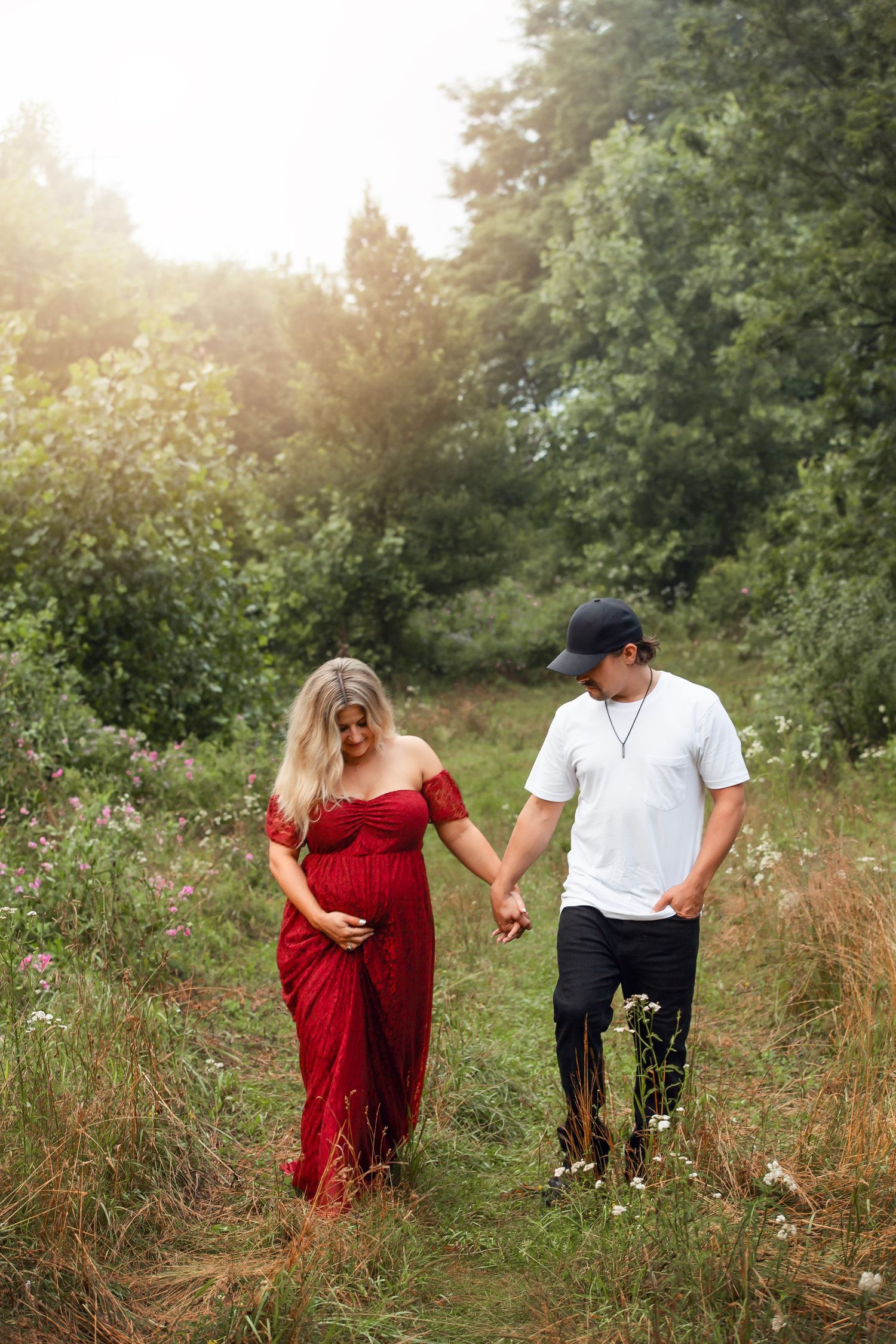 A couple in a meadow walking through tall grass at sunset, with her wearing a flowing red dress.