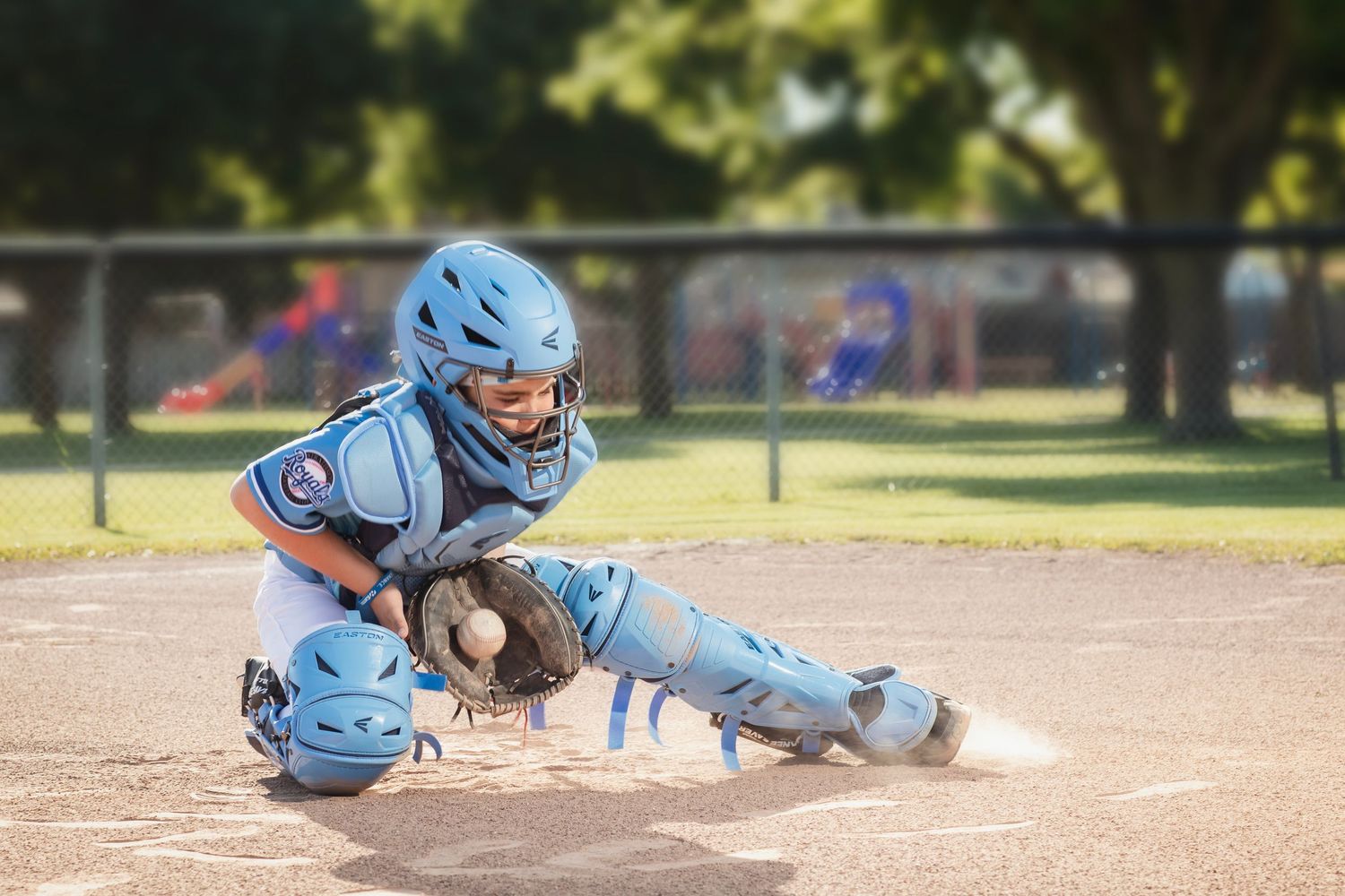 Youth baseball catcher in blue uniform blocking home plate during a game on a sunny day.