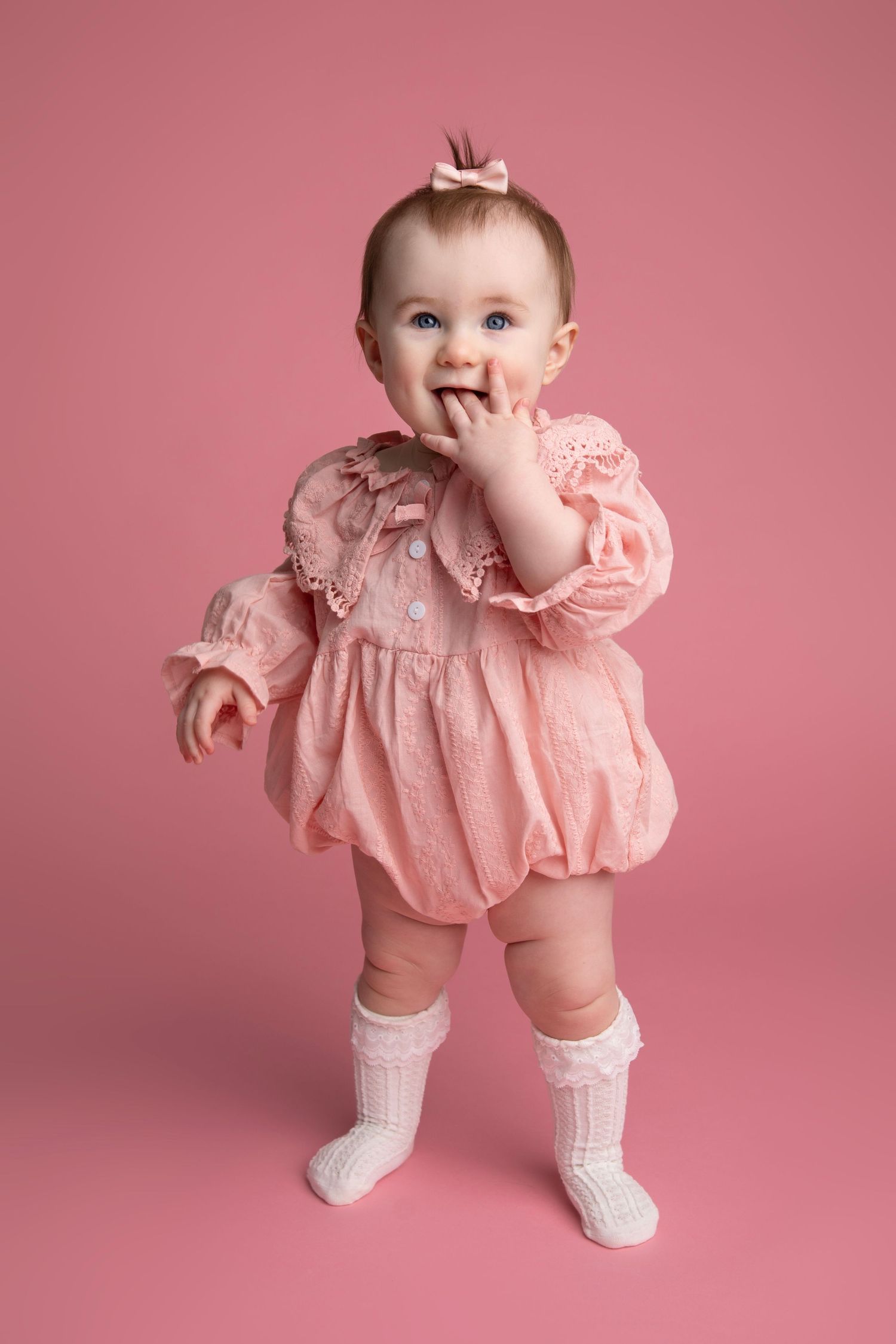 Baby in pink dress and white socks poses playfully against pink backdrop in series of sweet studio portraits.