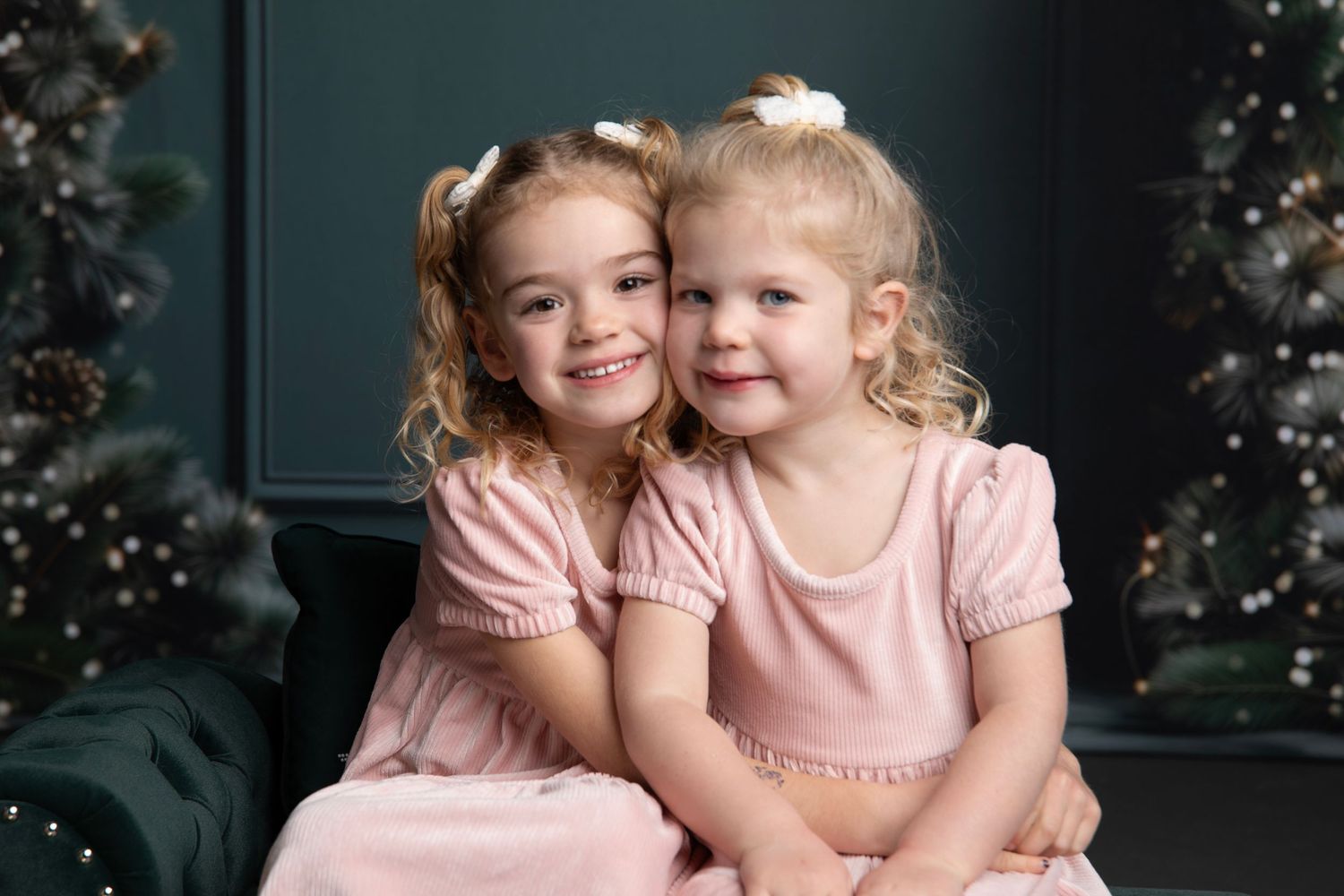 Two young children in pink dresses sit close together on a dark green couch with holiday decor in the background.